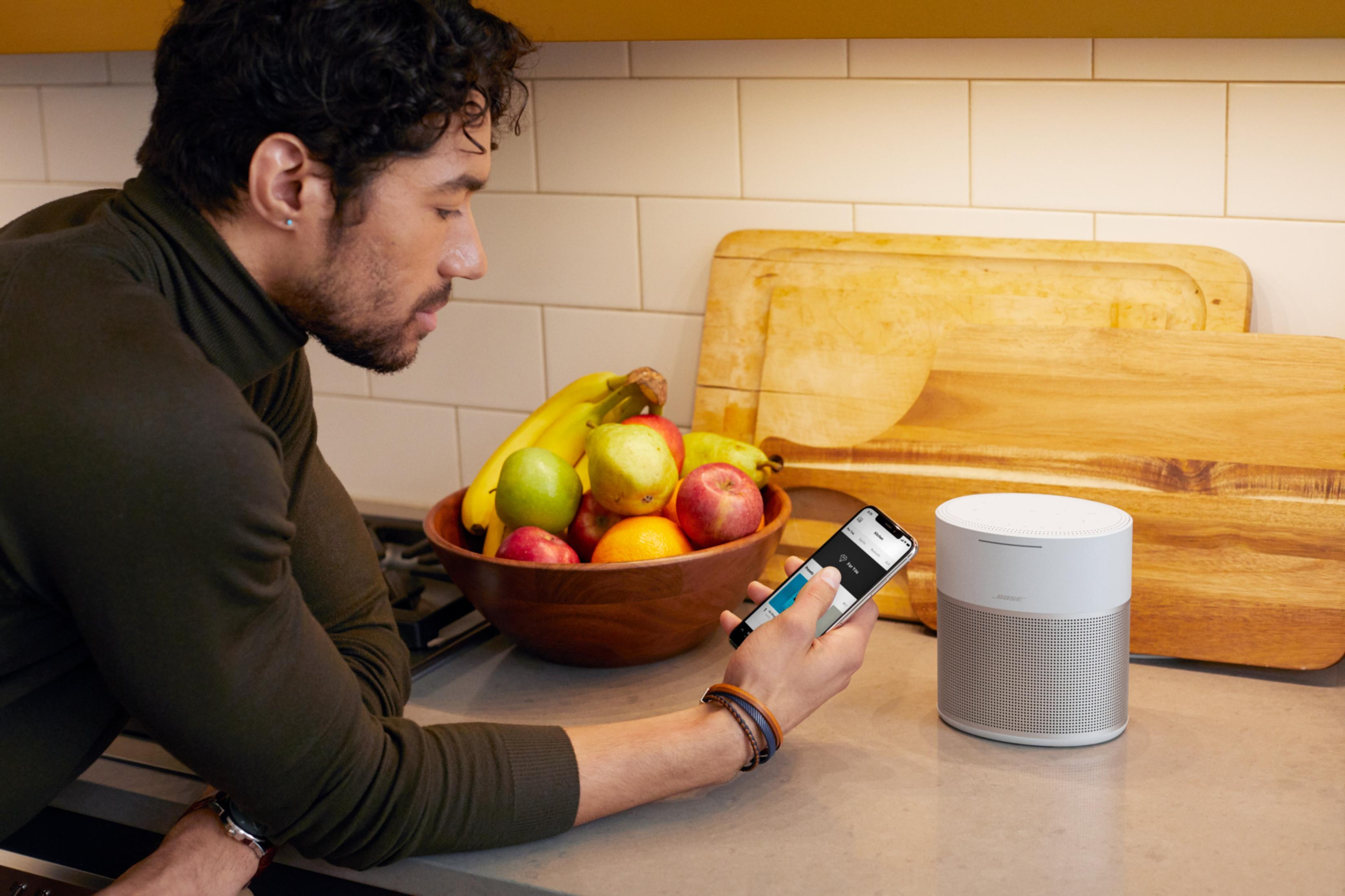 Ein Mann benutzt ein Telefon mit einem Smart Speaker, Obstschale auf einer Küchentheke.