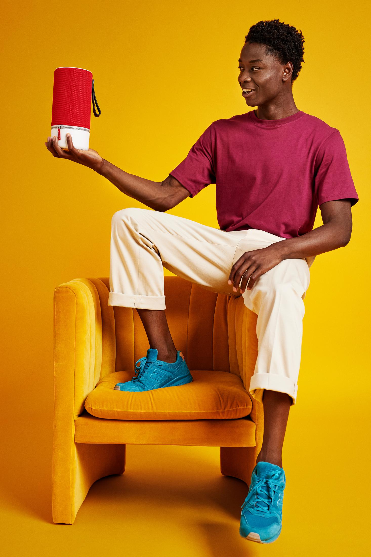 Młody czarnoskóry mężczyzna trzyma czerwono-biały głośnik, siedząc na żółtym krześle.
