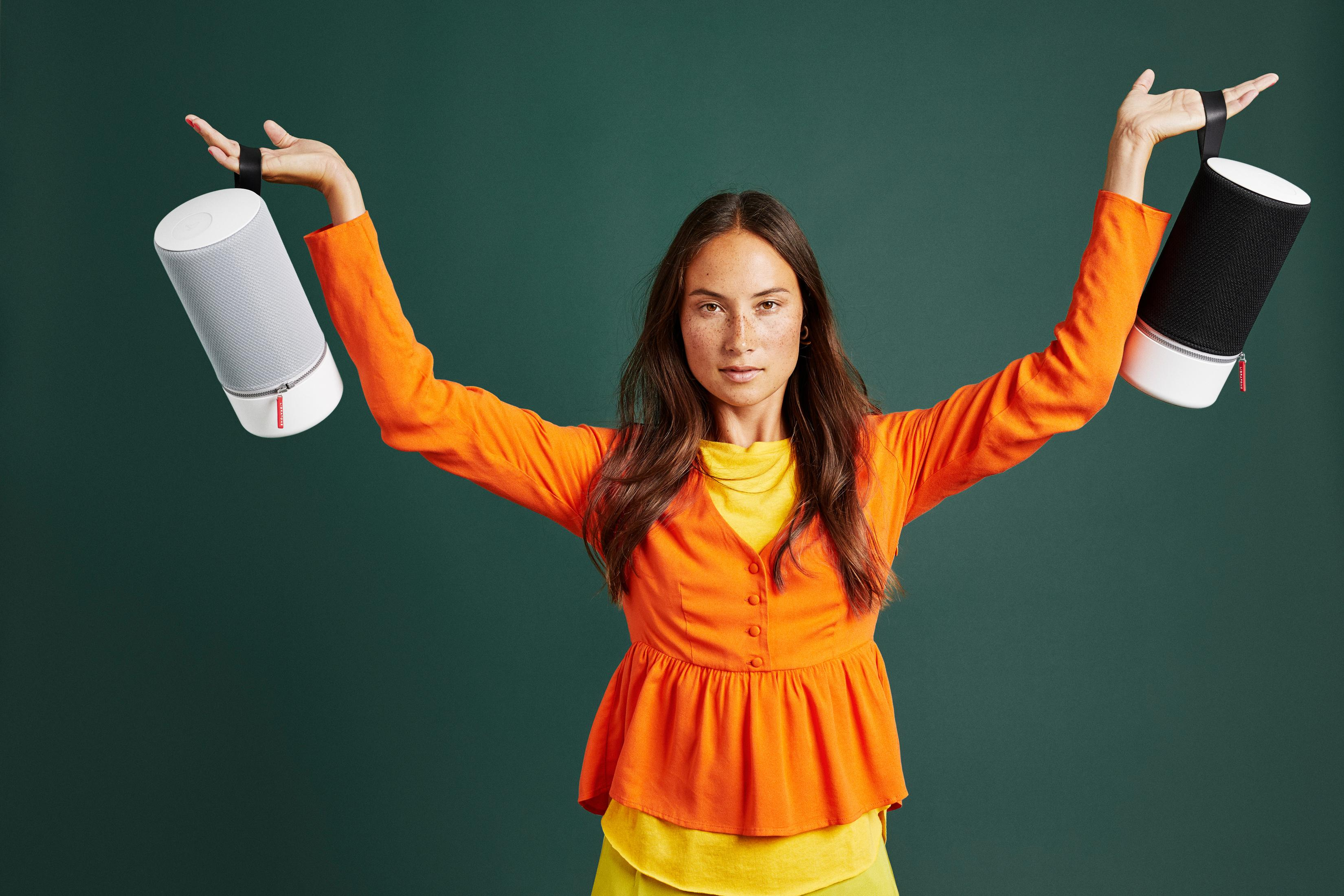 Kobieta trzyma dwa cylindryczne głośniki, szary i czarny, na tle turkusowego zielonego.
