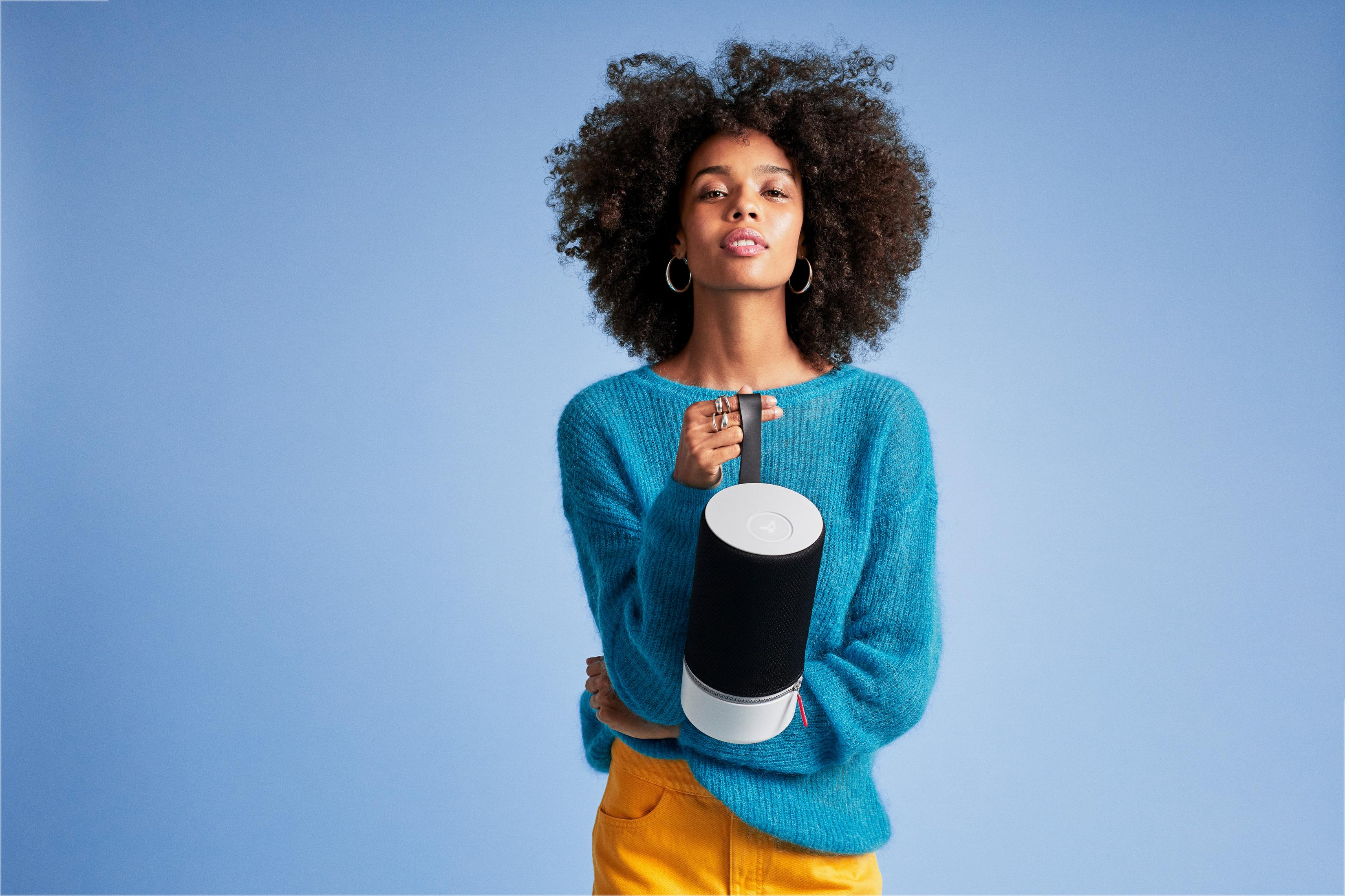 Kobieta z afro trzyma czarno-biały głośnik. Ma na sobie niebieski sweter i żółte spodnie na niebieskim tle.