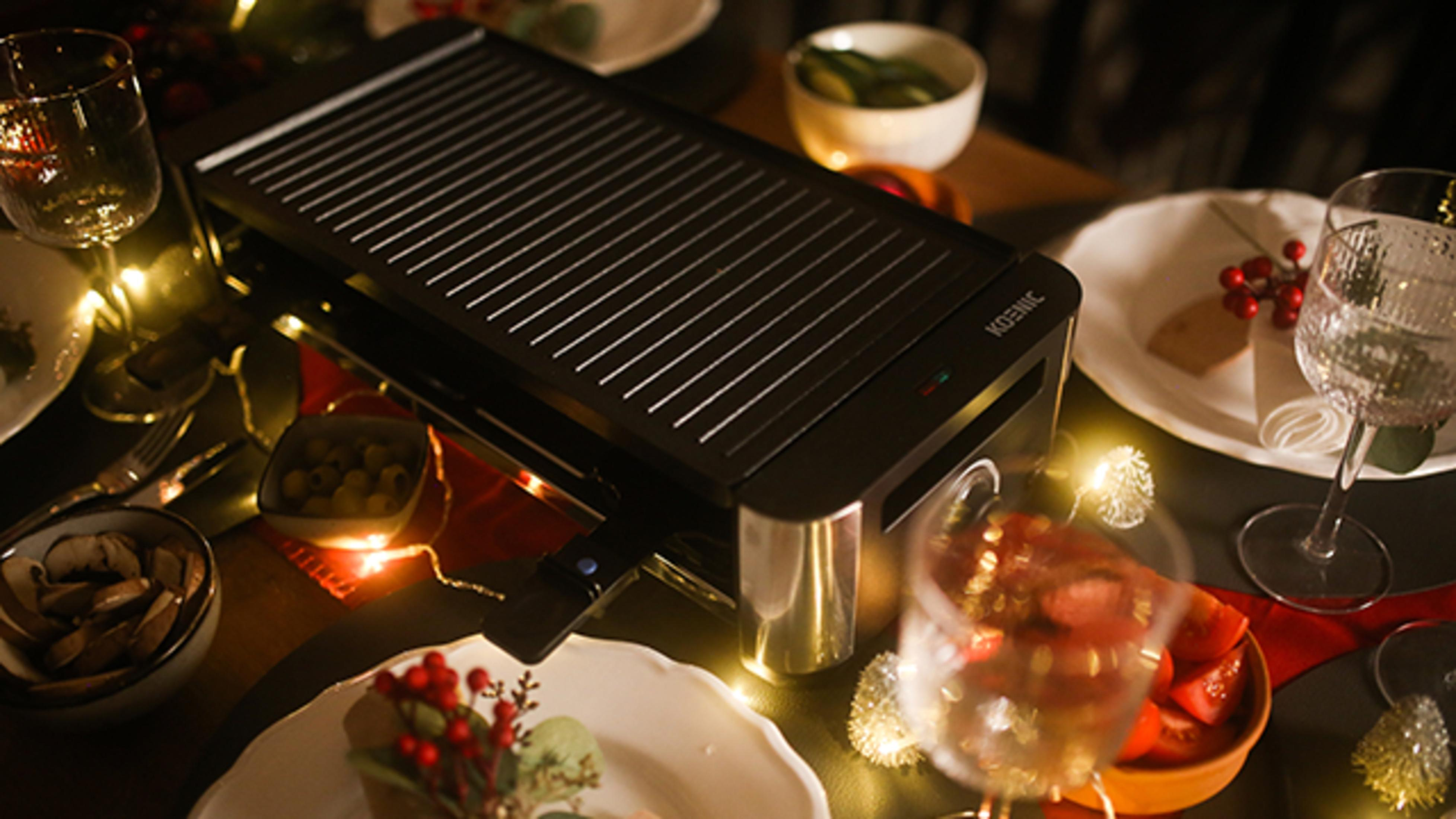 Ein schwarzer Koenic Raclette-Grill auf einem Tisch, gedeckt mit Essen, Gläsern und Lichtern.