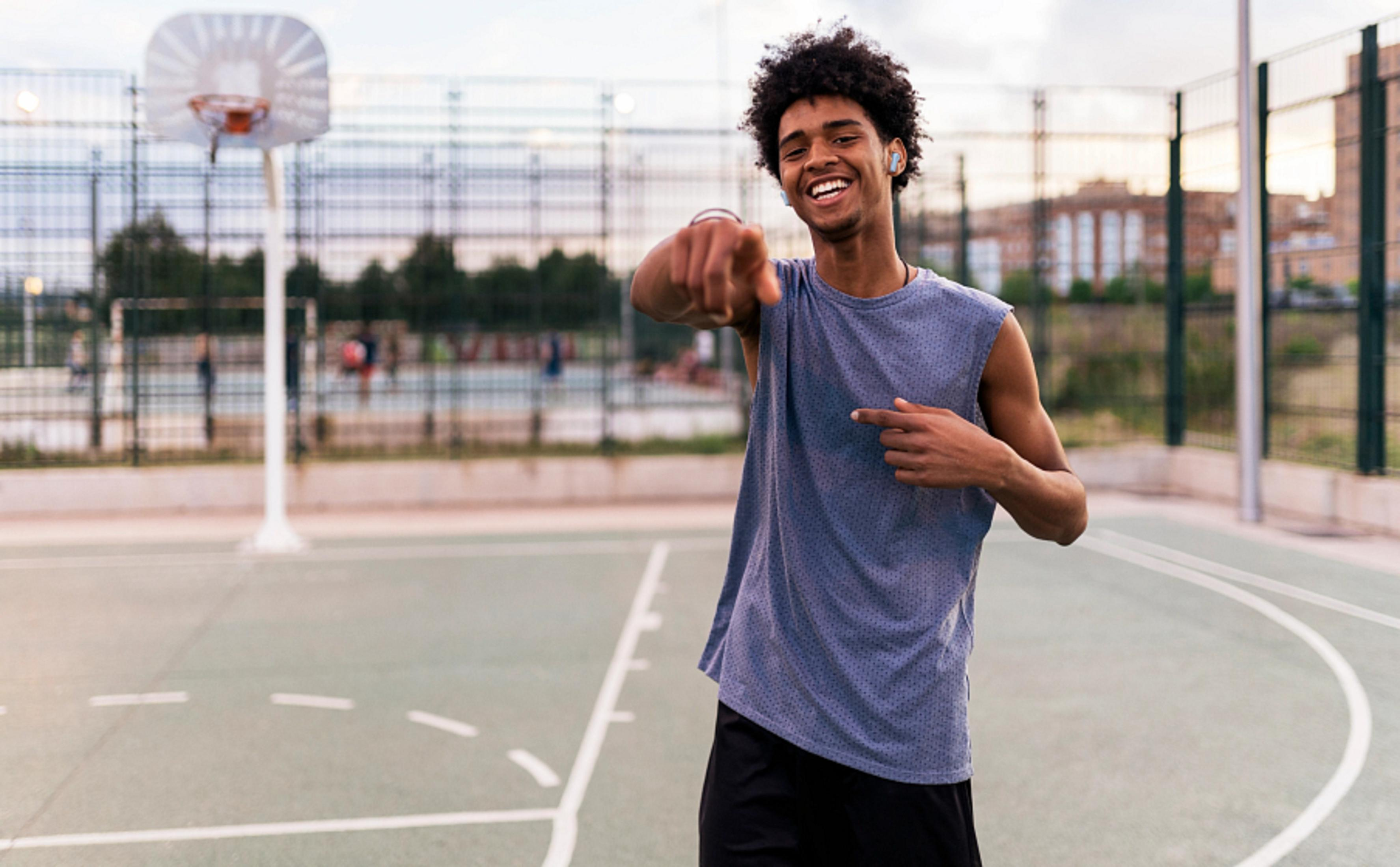 Młody czarny mężczyzna uśmiechnięty, wskazujący, z słuchawkami, na boisku do koszykówki.