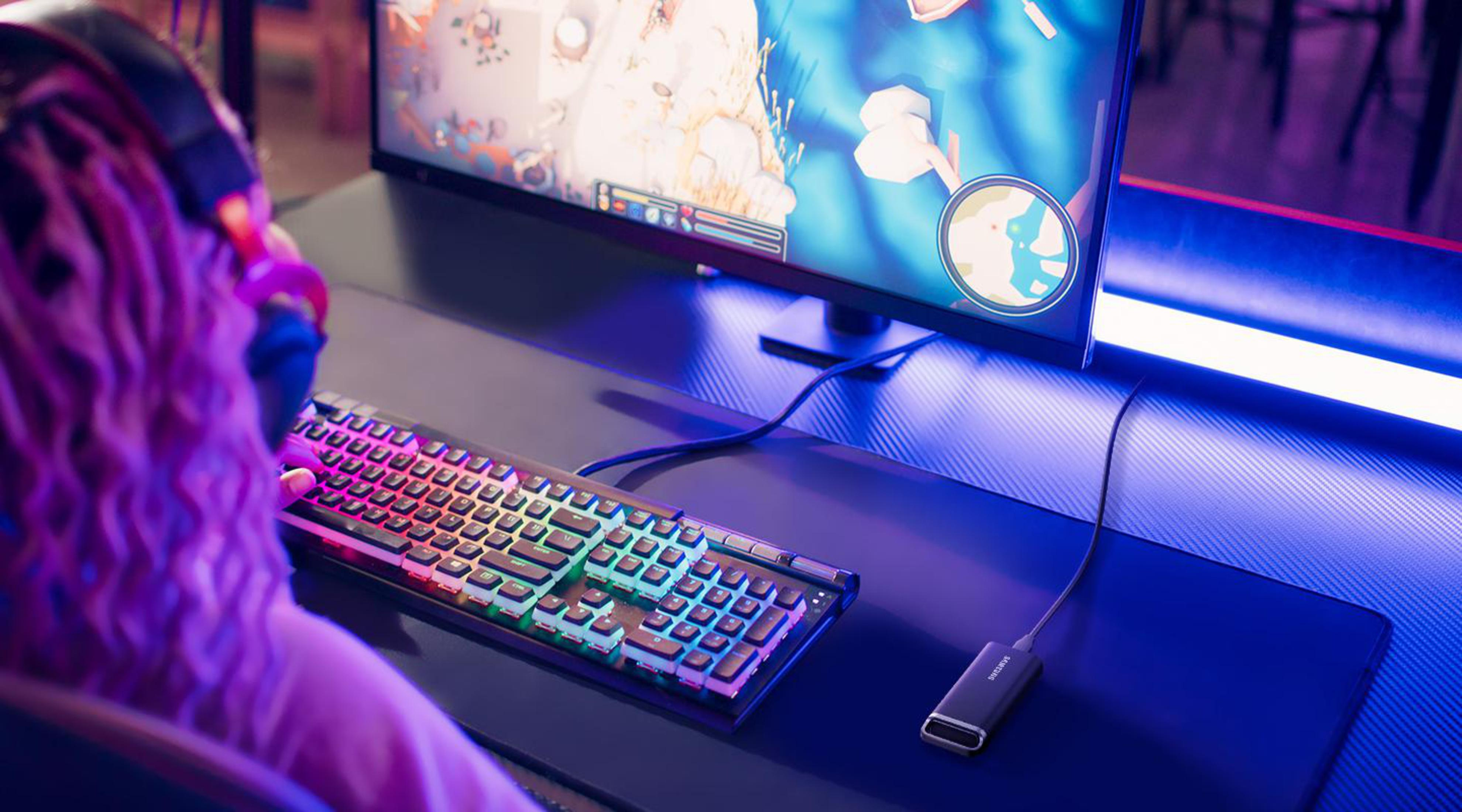 Osoba grająca w grę na komputerze z kolorową klawiaturą i monitorem na biurku.