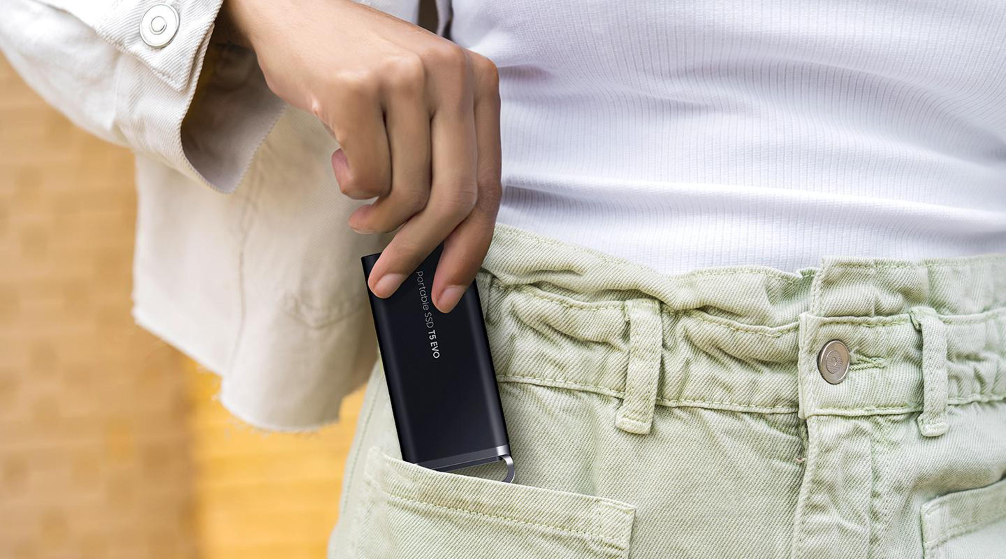 Une main sort un SSD portable noir d'une poche kaki. Le SSD a un accent argenté.