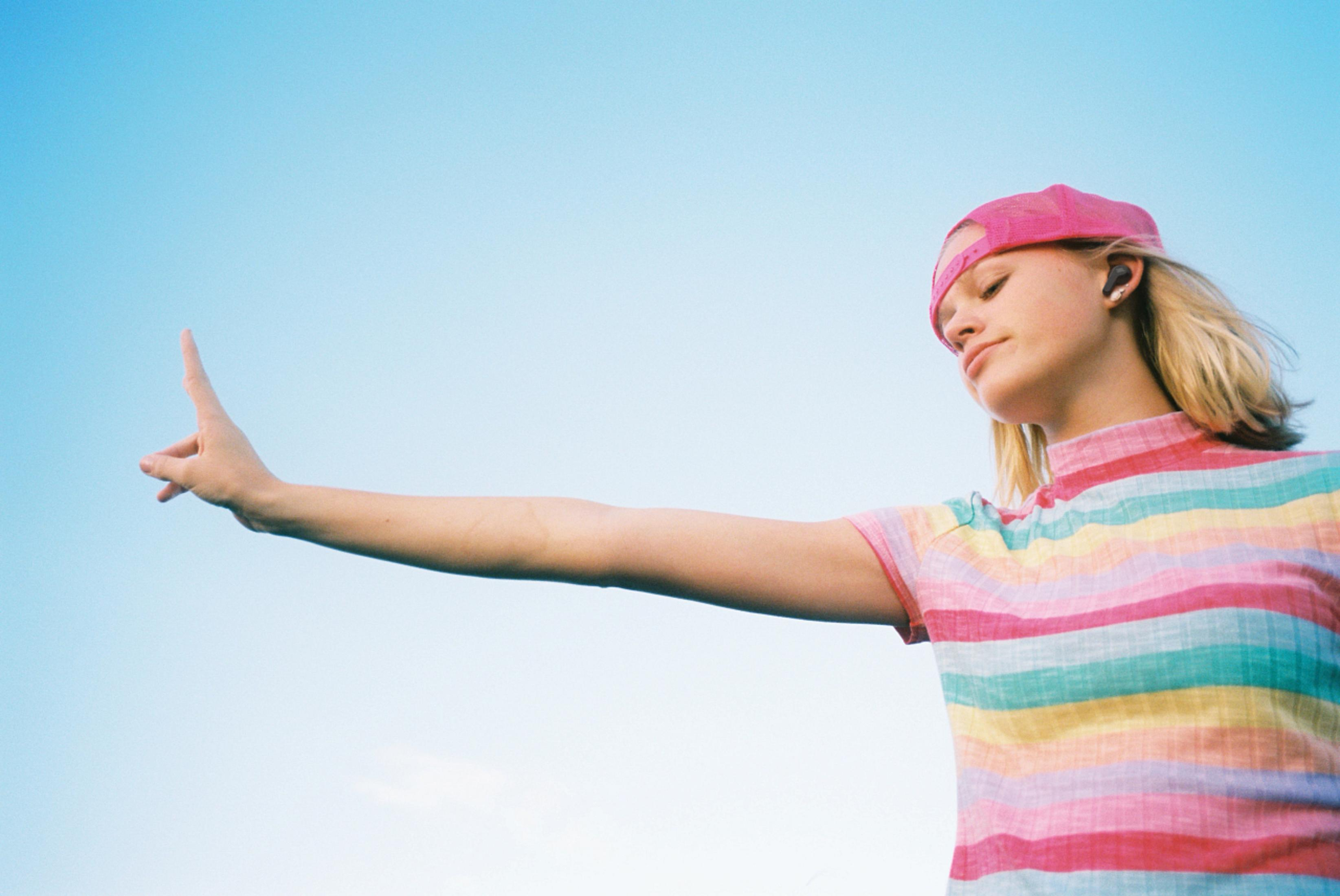 Junge Frau mit rosa Mütze und Ohrhörern gestikuliert zum Himmel, blauer Hintergrund.