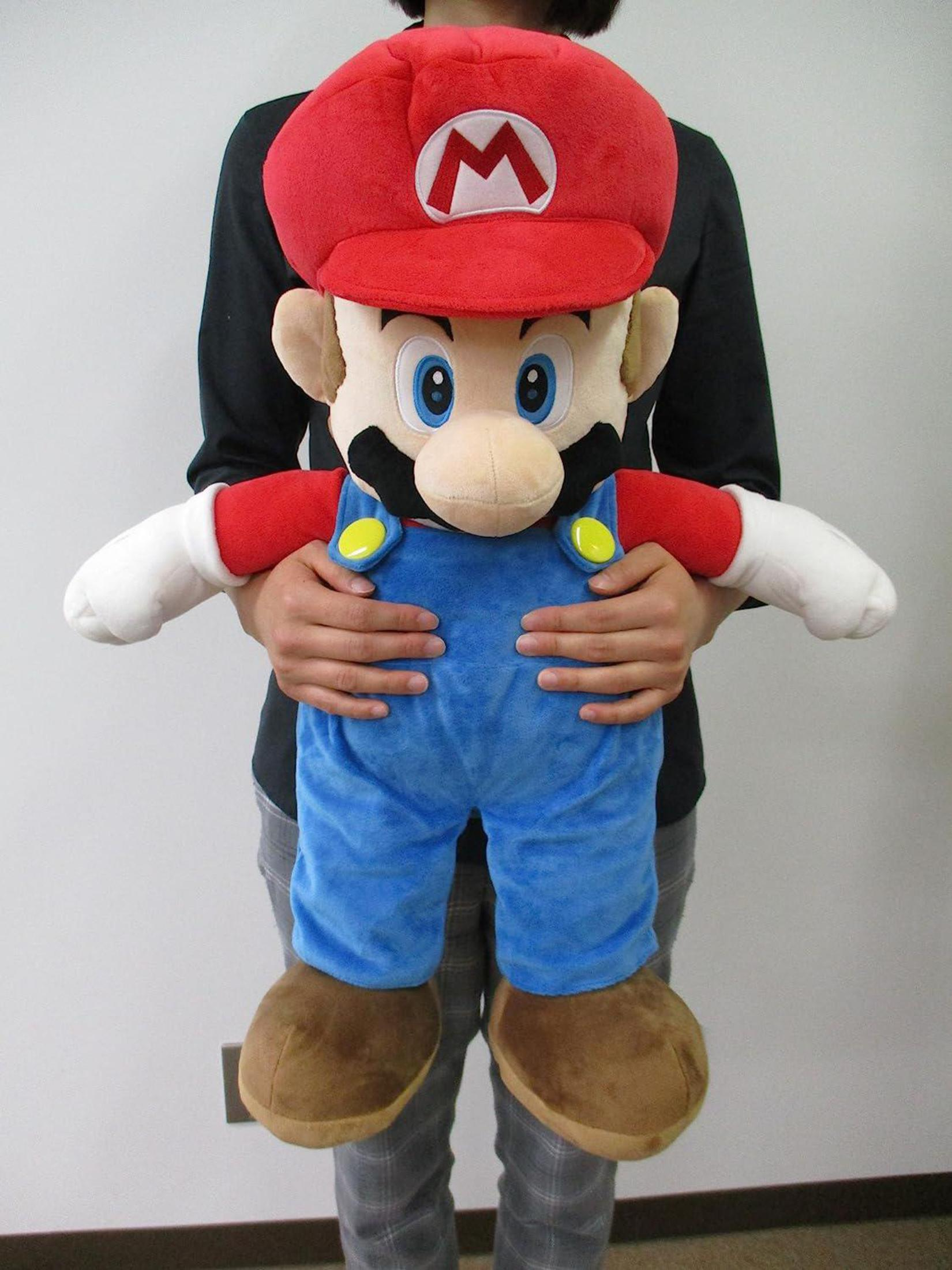 Una persona tiene in mano un grande peluche di Mario. Mario indossa cappello rosso, salopette blu e scarpe marroni.