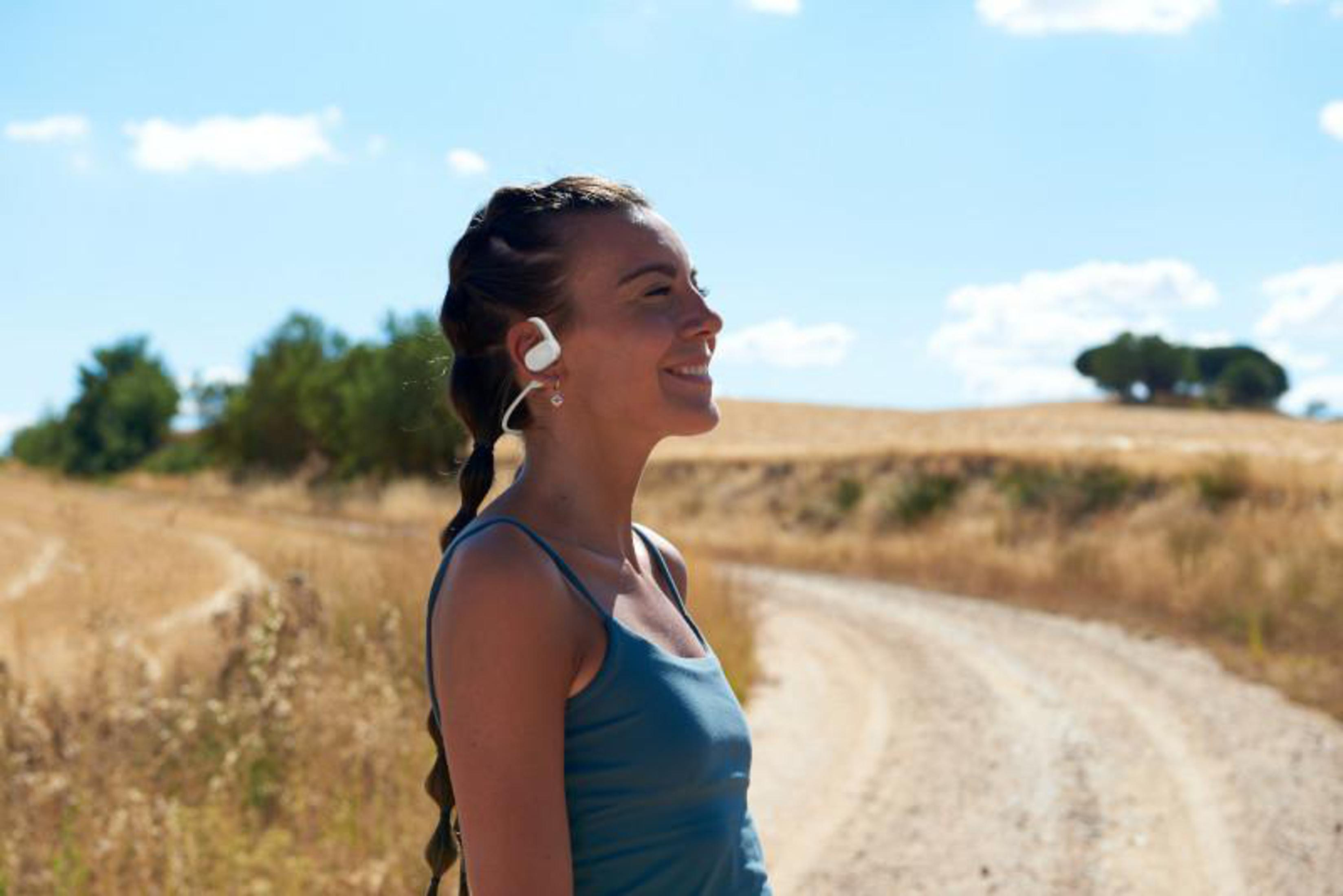 Mujer corriendo con auriculares. Sonríe y lleva una camiseta sin mangas azul. Campo al fondo.
