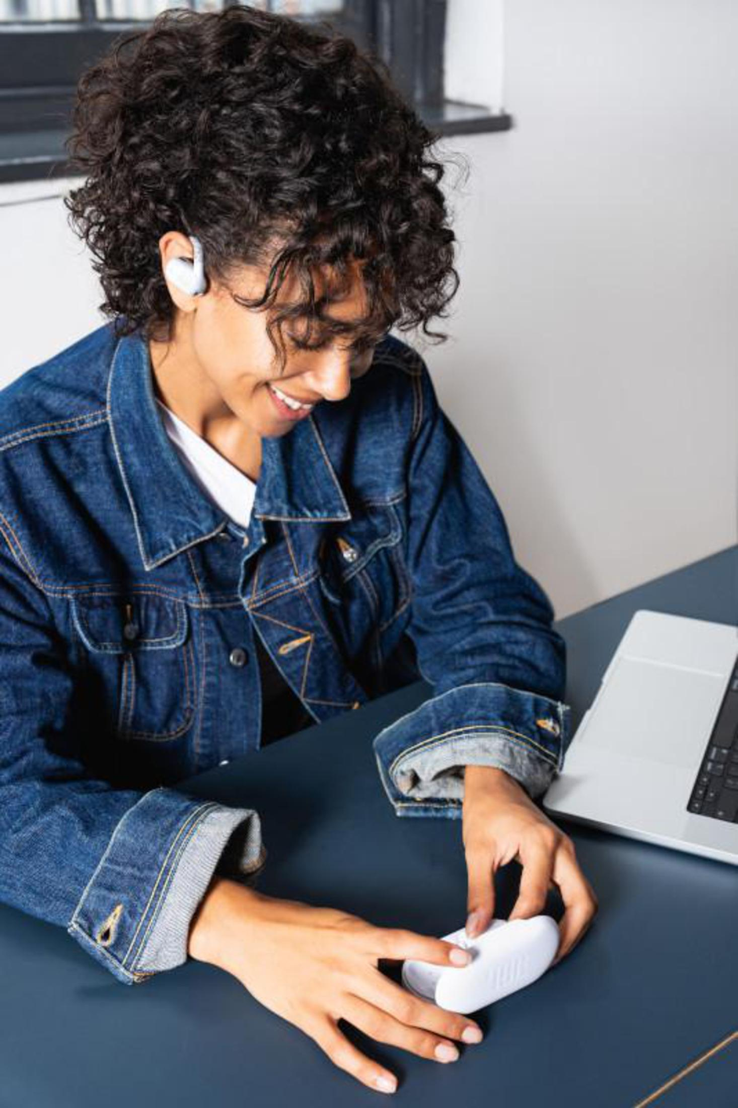 Mujer con pelo rizado, chaqueta de mezclilla, usando auriculares inalámbricos blancos, sonriendo.