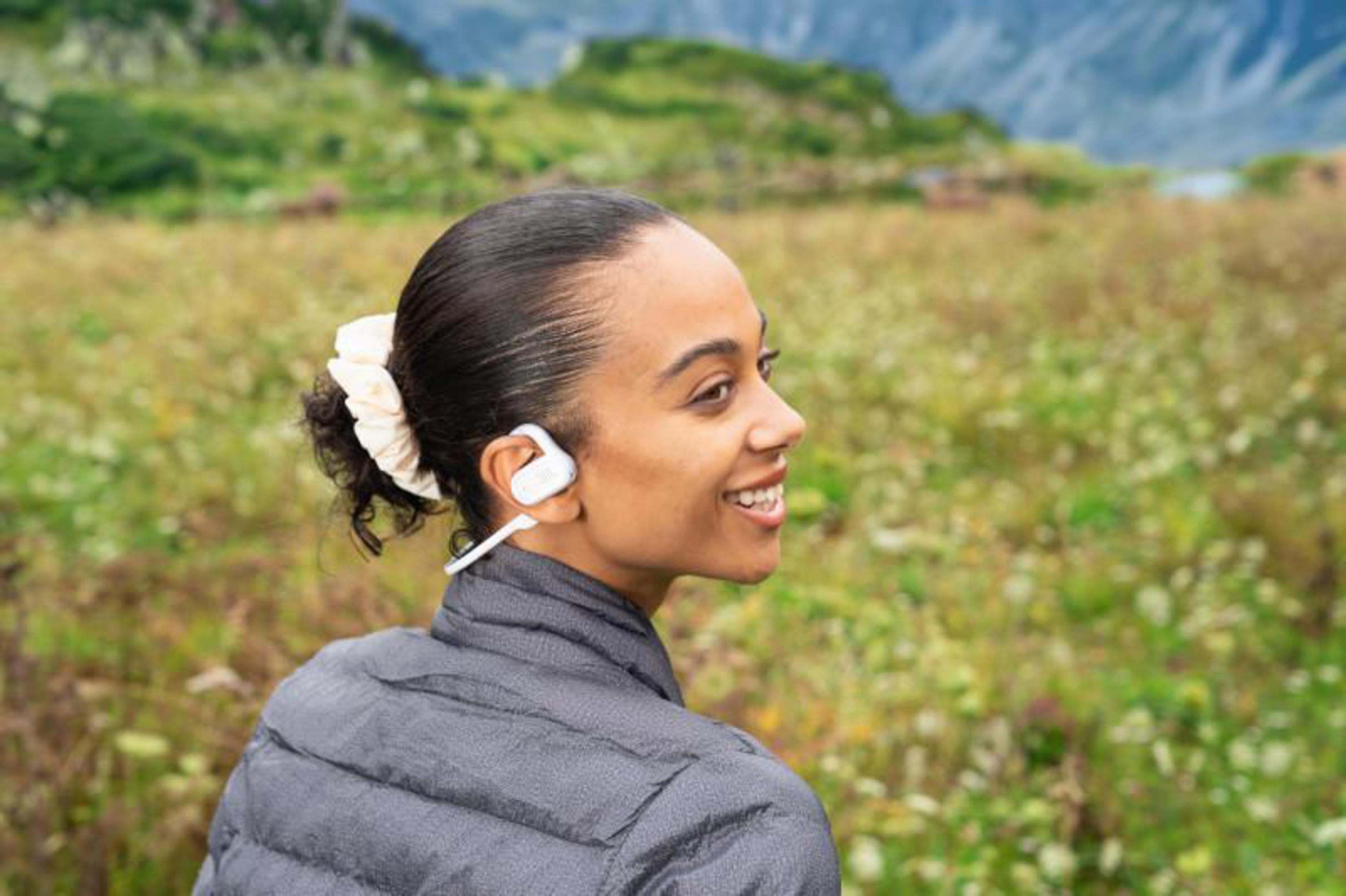 Mujer sonriente con auricular blanco y chaqueta acolchada gris. Campo verde y montañas al fondo.