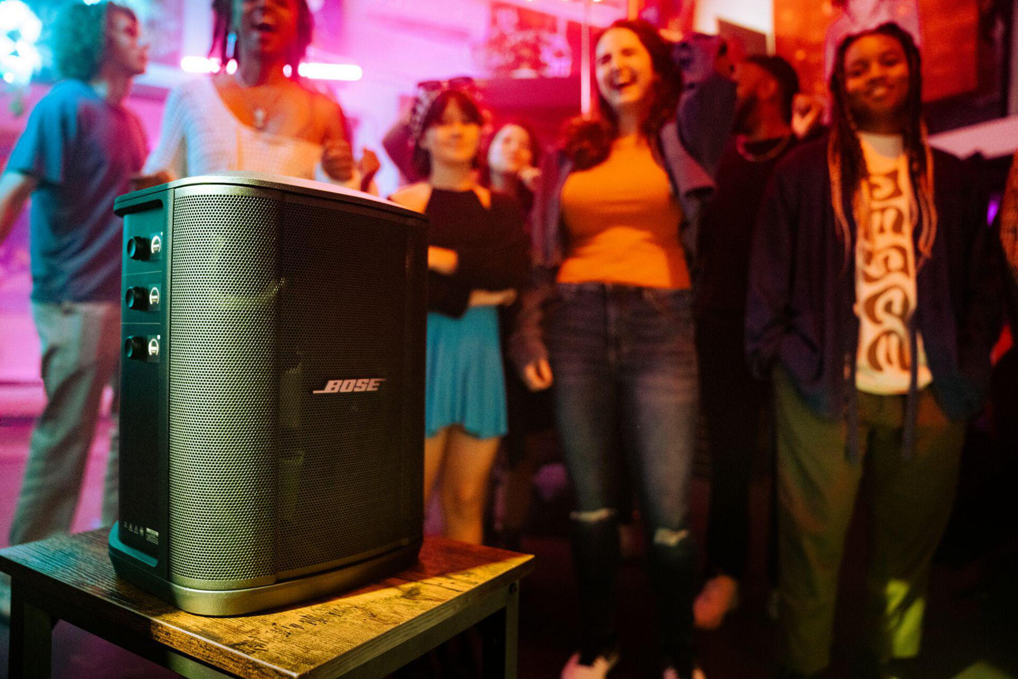 Un altavoz Bose en una mesa con gente al fondo. El altavoz es negro y la gente está borrosa.