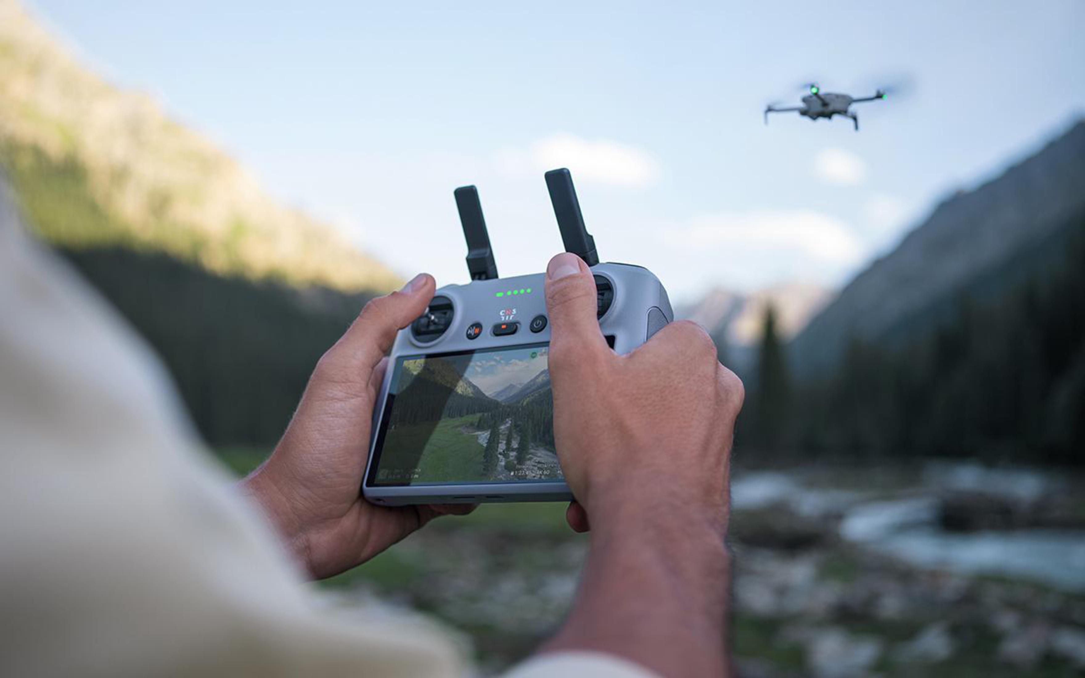 Dłonie trzymające kontroler drona. Dron leci w tle nad górskim krajobrazem.