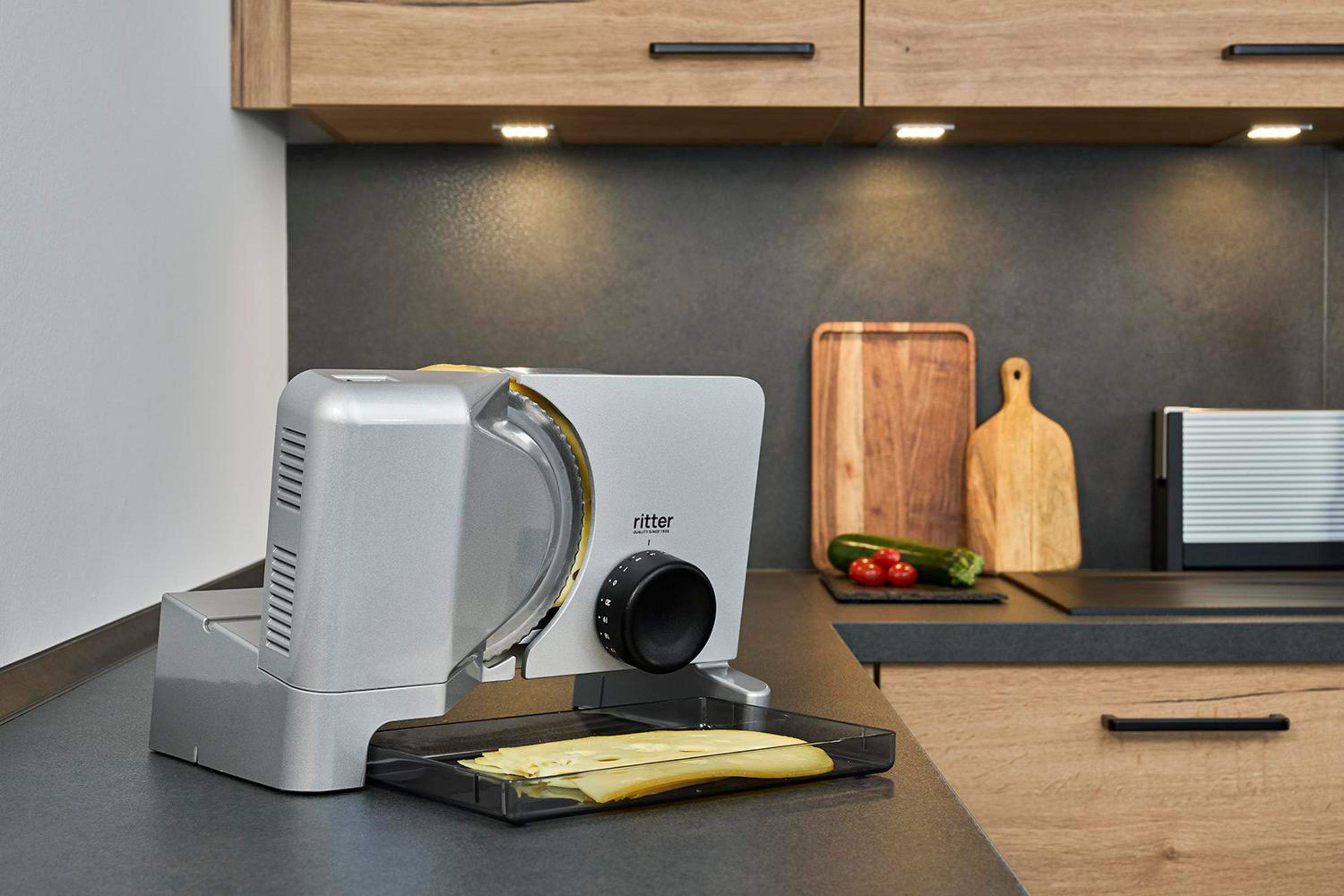 Srebrna krajalnica do żywności z plastrami sera na tacy, ustawiona na blacie w kuchni.
