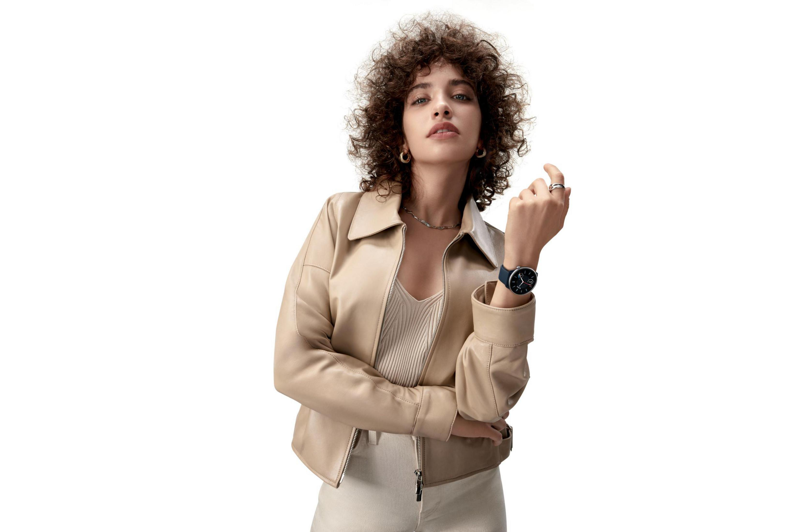 Kobieta z kręconymi włosami w beżowej kurtce i zegarkiem. Stoi na białym tle.