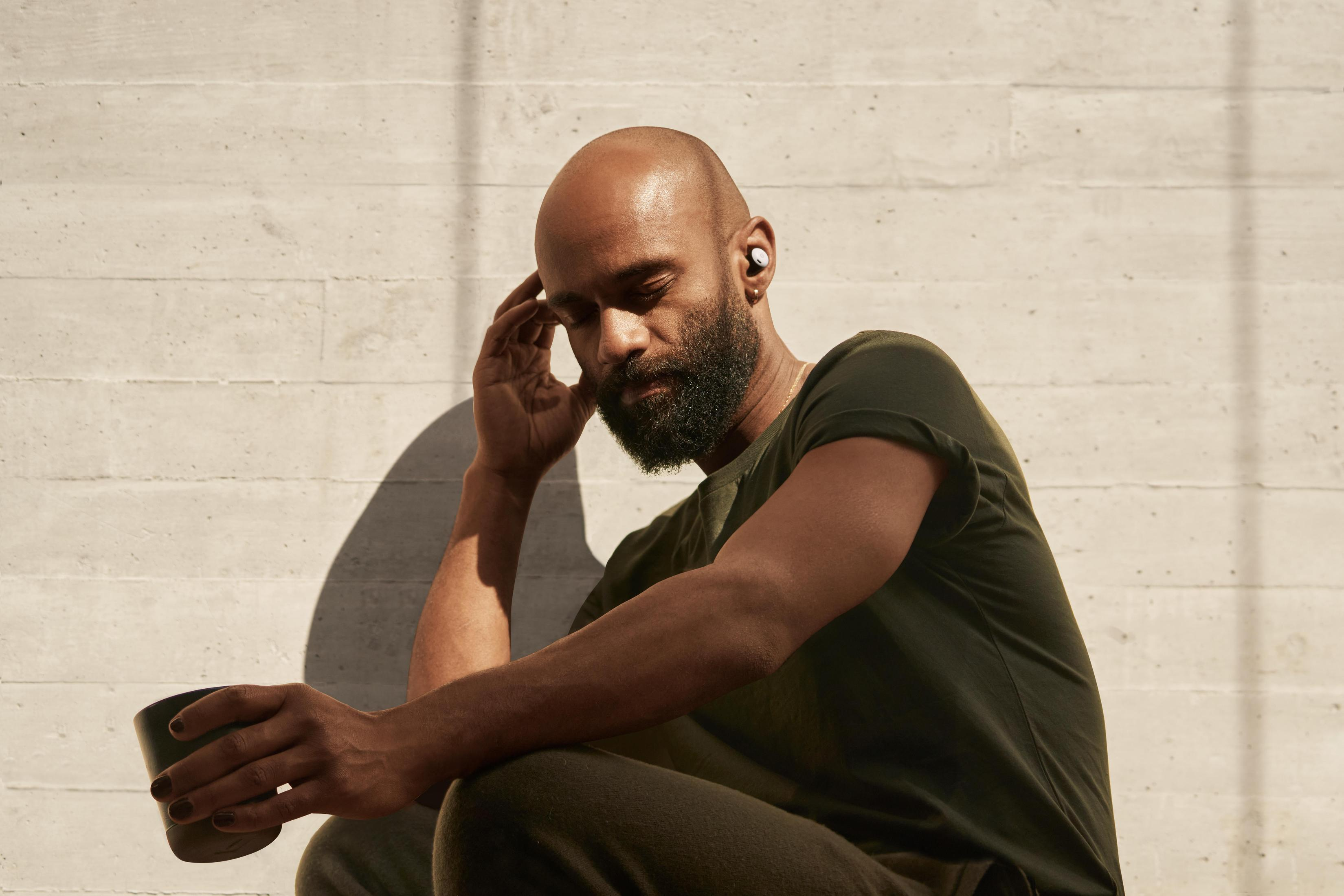 Mężczyzna z brodą w słuchawkach, trzymający czarny kubek, oparty o ścianę.