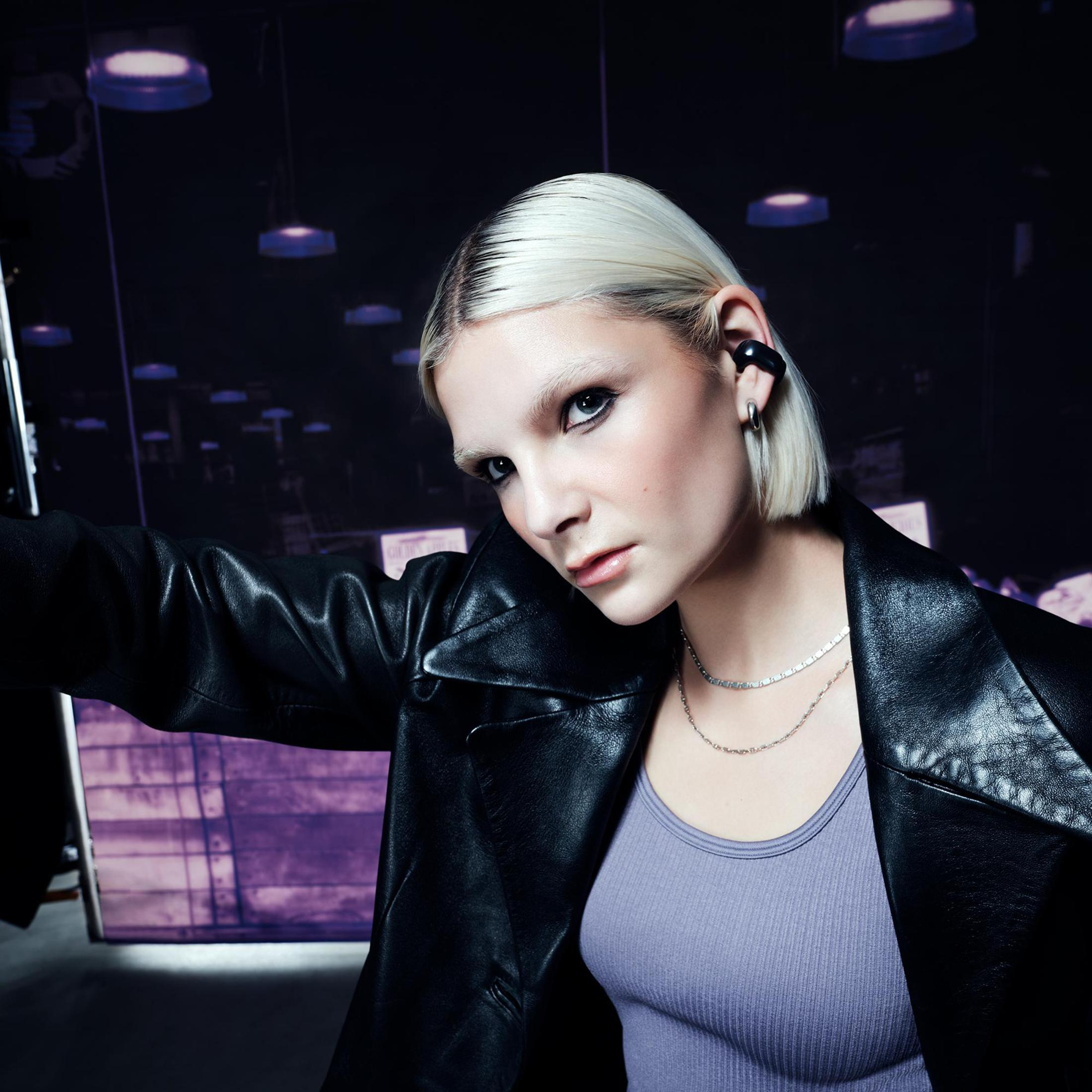 Mujer rubia con chaqueta de cuero negro y top morado, con auriculares y joyas de plata.