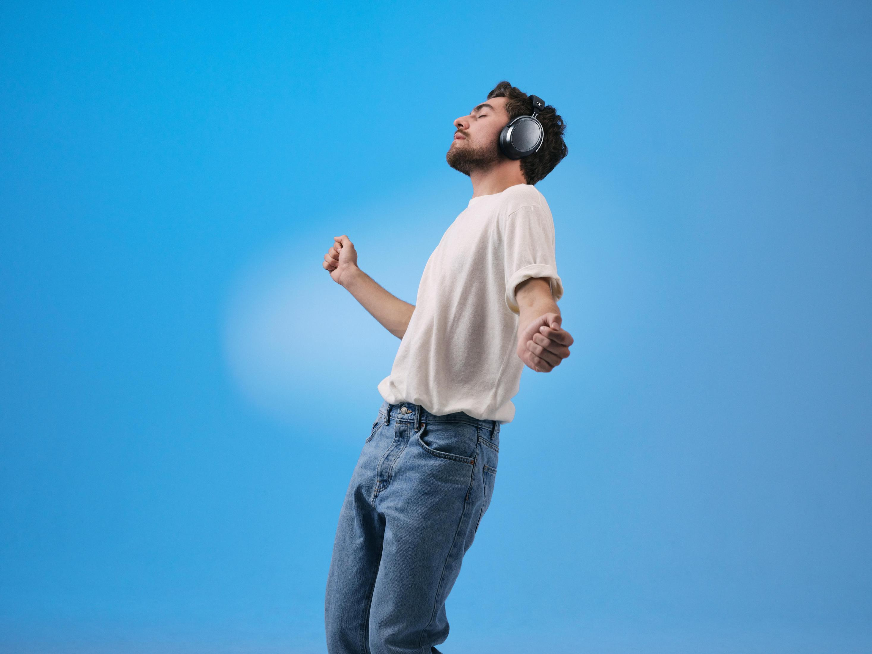 Mężczyzna z słuchawkami tańczy, oczy zamknięte, ramiona w górze, na niebieskim tle.