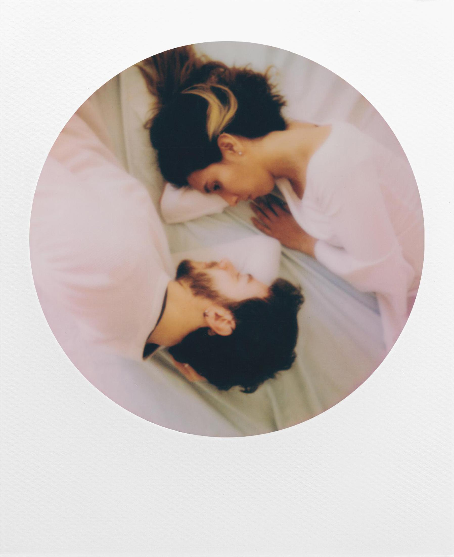 Polaroid-Foto eines Paares, das in einem kreisförmigen Rahmen auf einem Bett liegt. Das Bild ist weich und warm.
