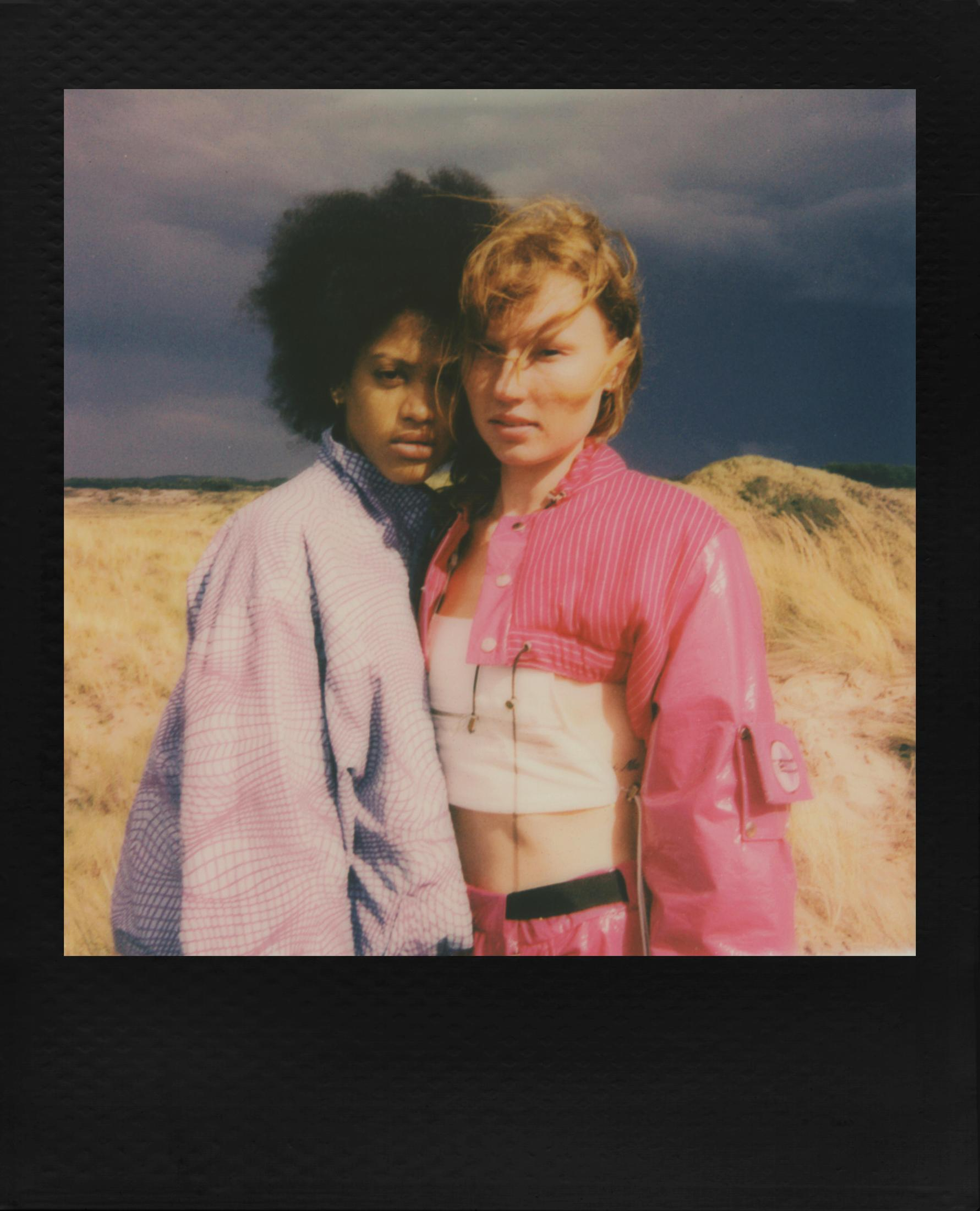 Dos mujeres frente a un paisaje. Una lleva una chaqueta rosa, la otra una blusa estampada.