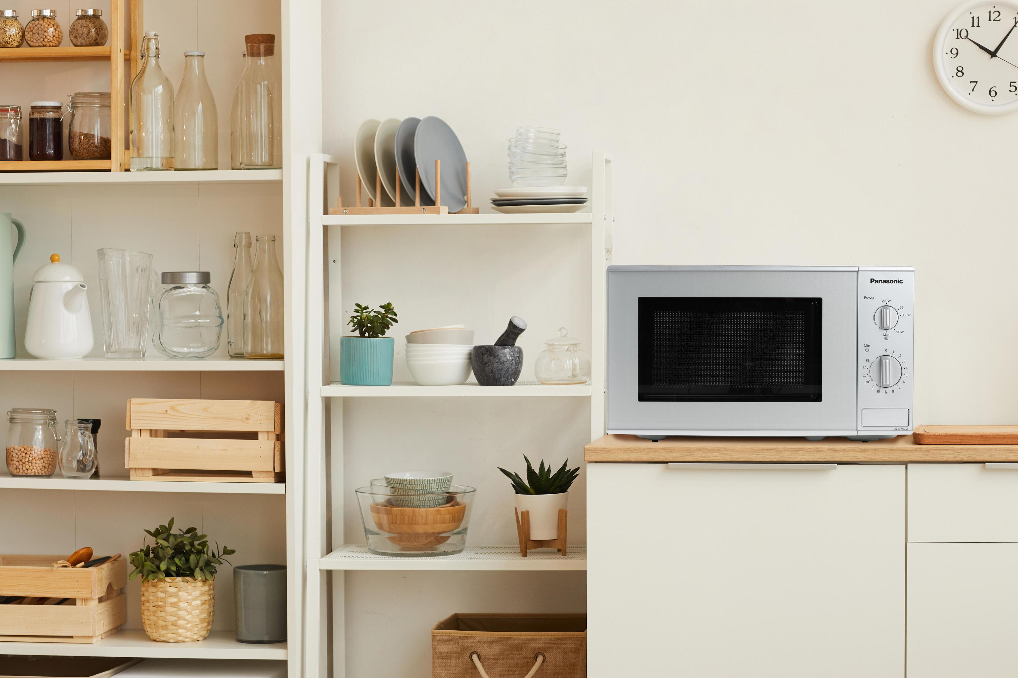 Srebrna kuchenka mikrofalowa na białym blacie w kuchni. Widoczne półki z naczyniami, słoikami i roślinami.