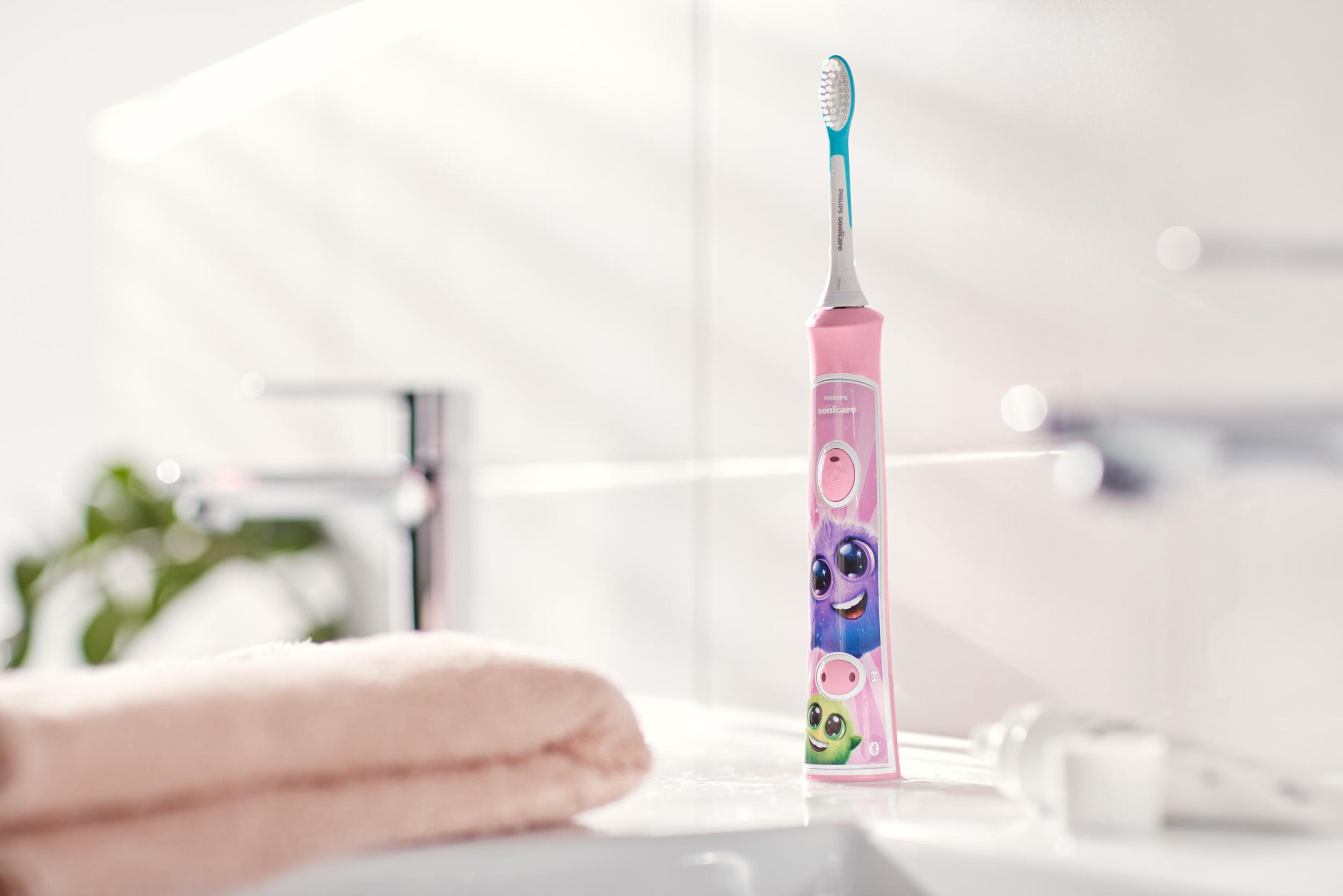 Cepillo de dientes rosa con personajes de dibujos animados, colocado sobre un lavabo blanco con una toalla rosa al fondo.