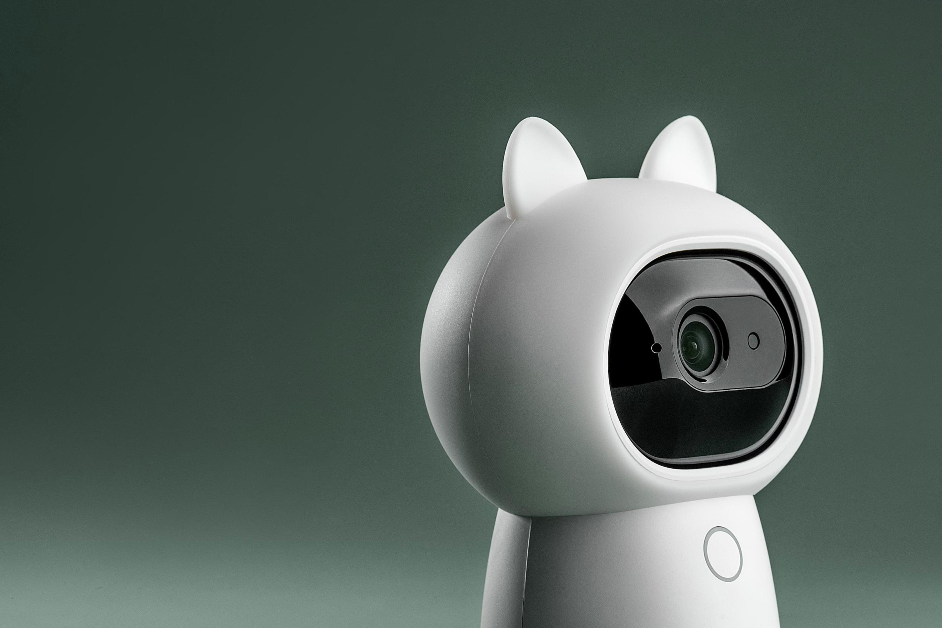 Biała kamera bezpieczeństwa w kształcie kota z czarnym obiektywem i dwoma uszami na zielonym tle.