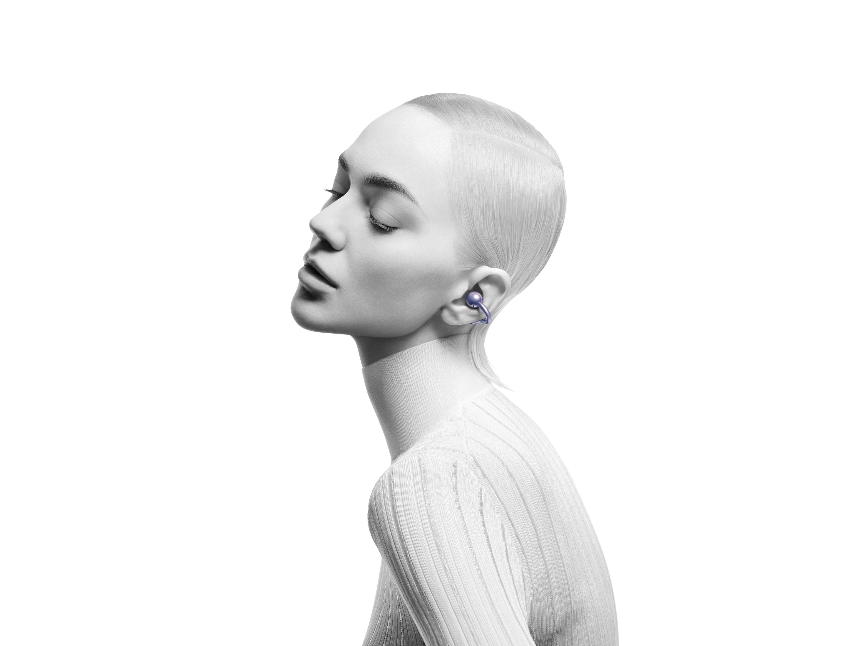 Kobieta z krótkimi włosami, nosząca słuchawkę w lewym uchu, zwrócona w lewo. Ma na sobie biały sweter z golfem.
