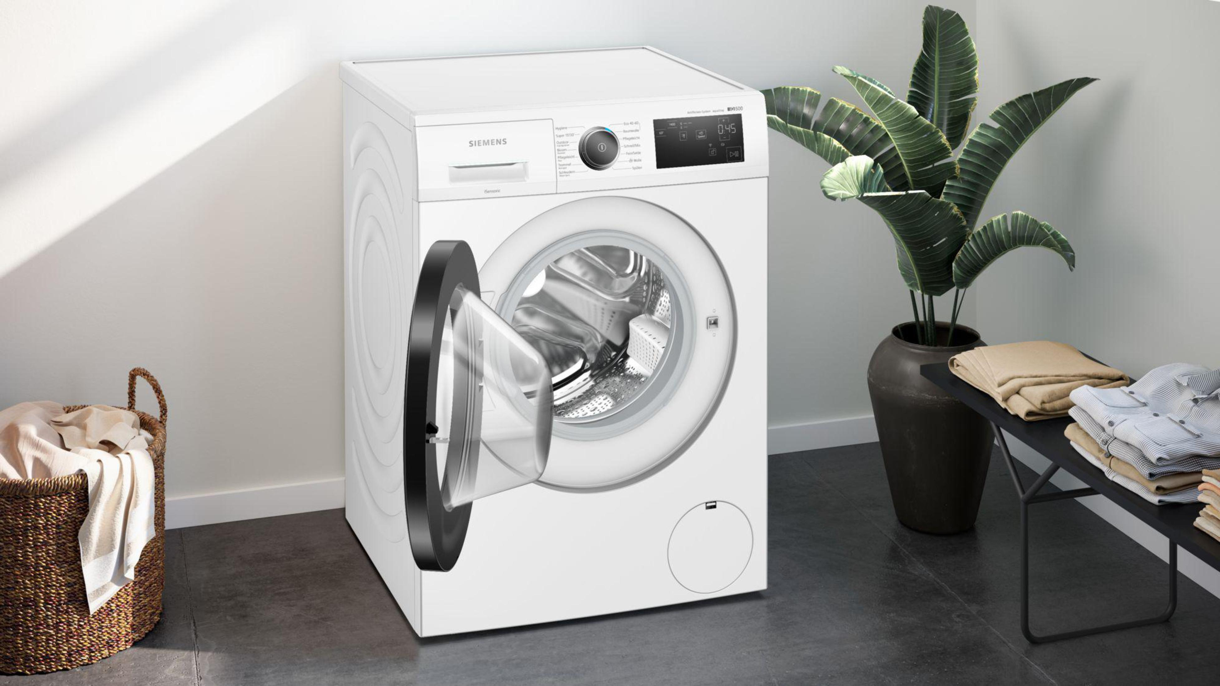 Weiße Waschmaschine mit offener Tür. Ein Korb, Pflanze und Kleidung daneben.