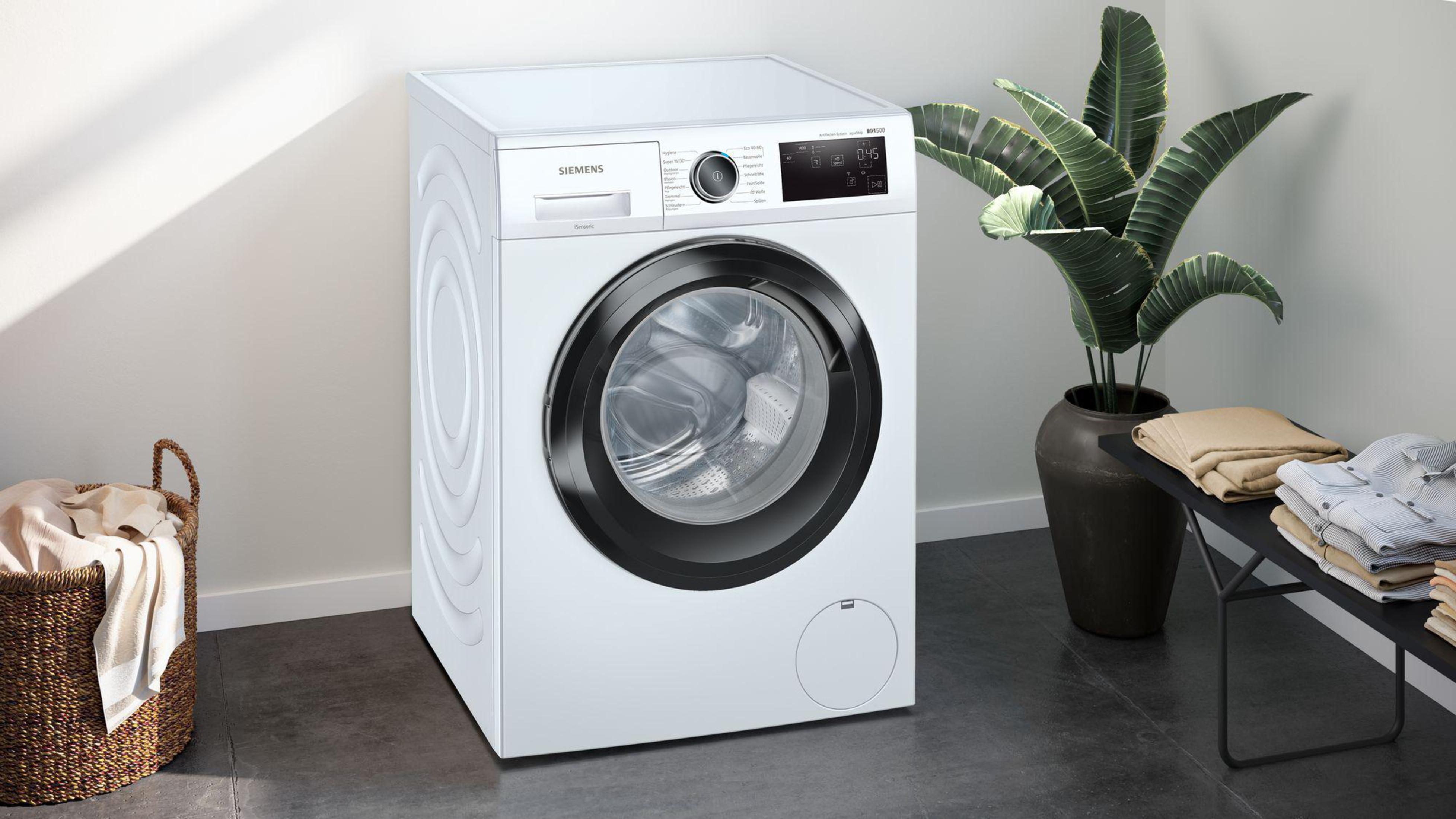 Eine weiße Siemens Waschmaschine mit schwarzen Details, neben einer Pflanze und einem Wäschekorb.