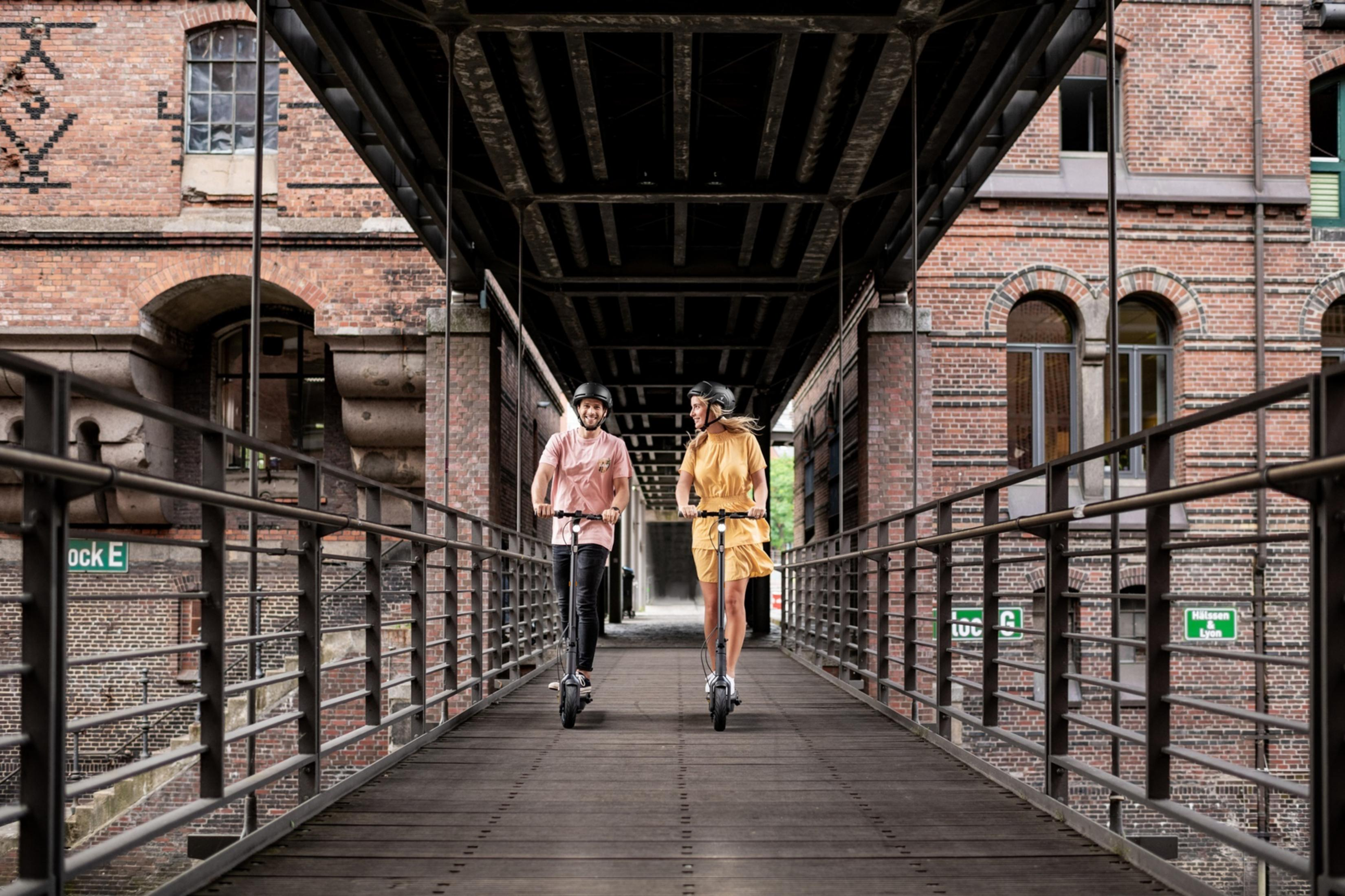 Zwei Personen fahren mit Elektrorollern auf einer Brücke, Backsteingebäude im Hintergrund.