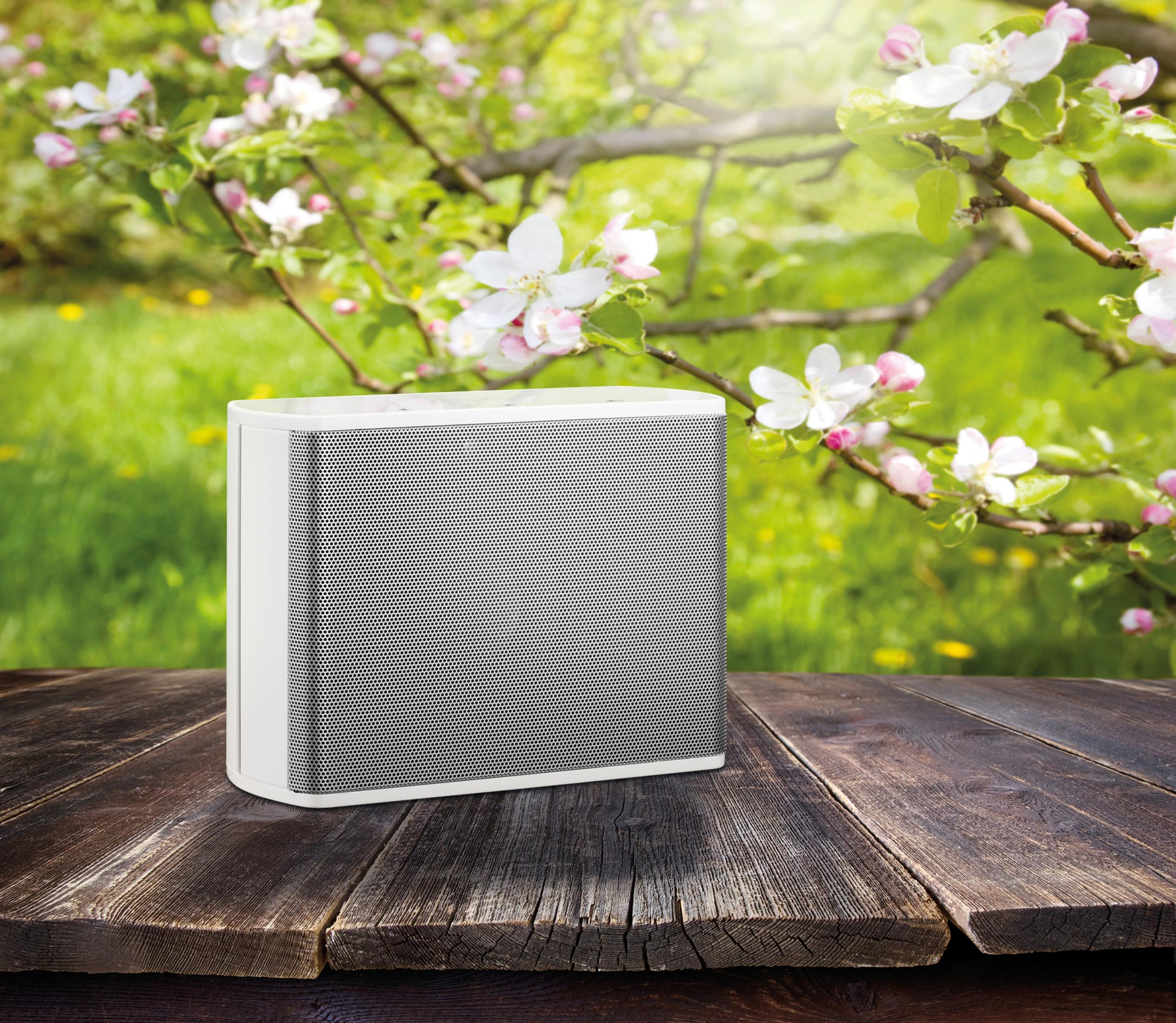 White speaker on a wooden table in front of a blurred green and white flowery background.