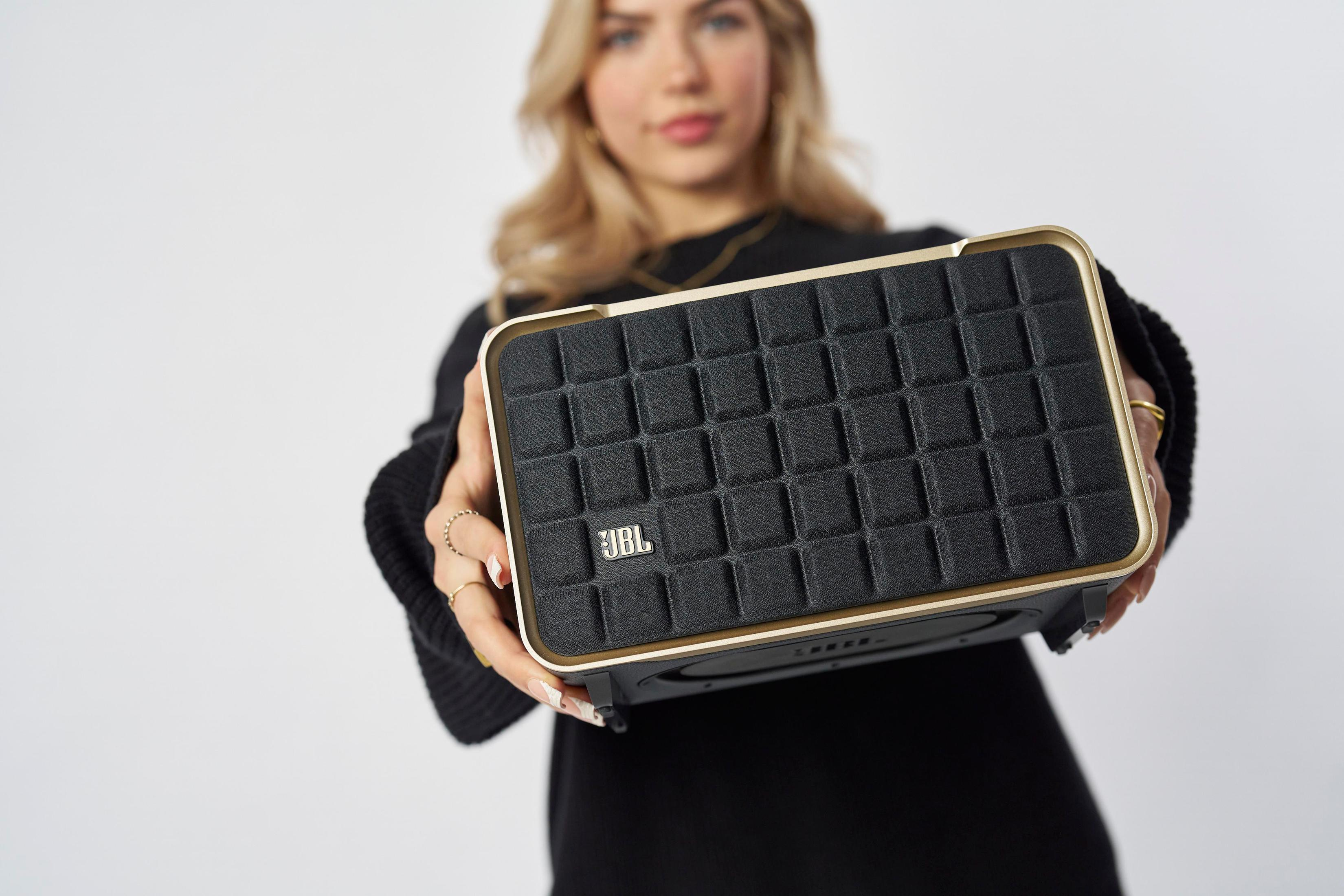 Kobieta trzyma czarny głośnik ze złotymi akcentami i logo JBL.