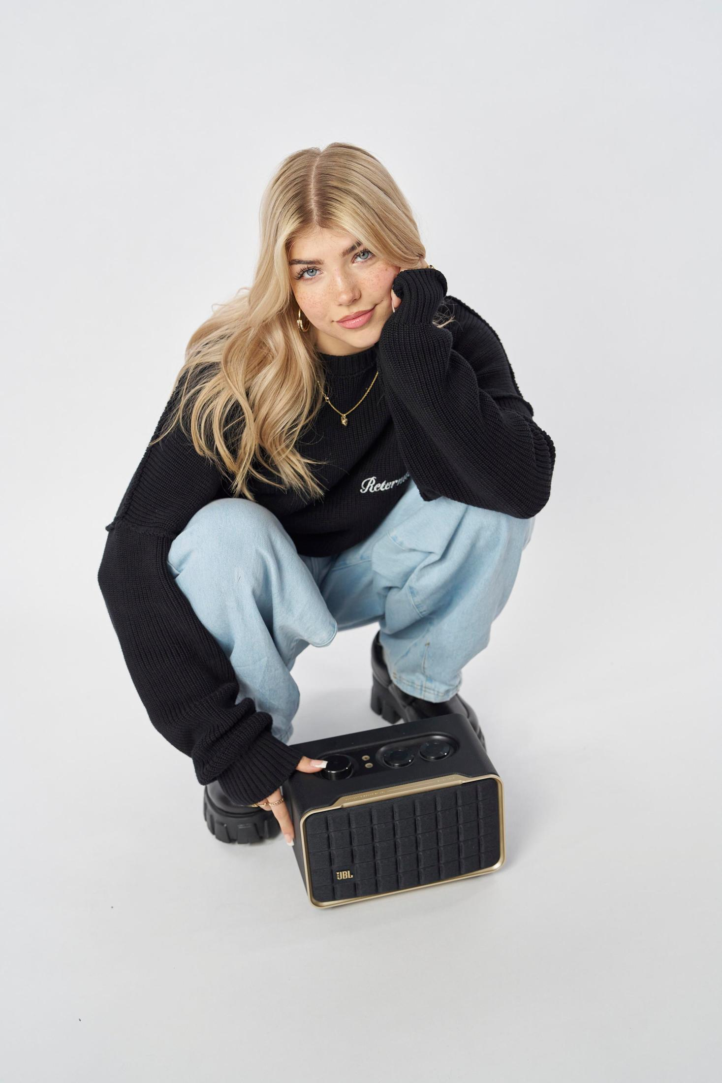 Kobieta w czarnym swetrze i niebieskich dżinsach kuca, trzymając głośnik JBL.