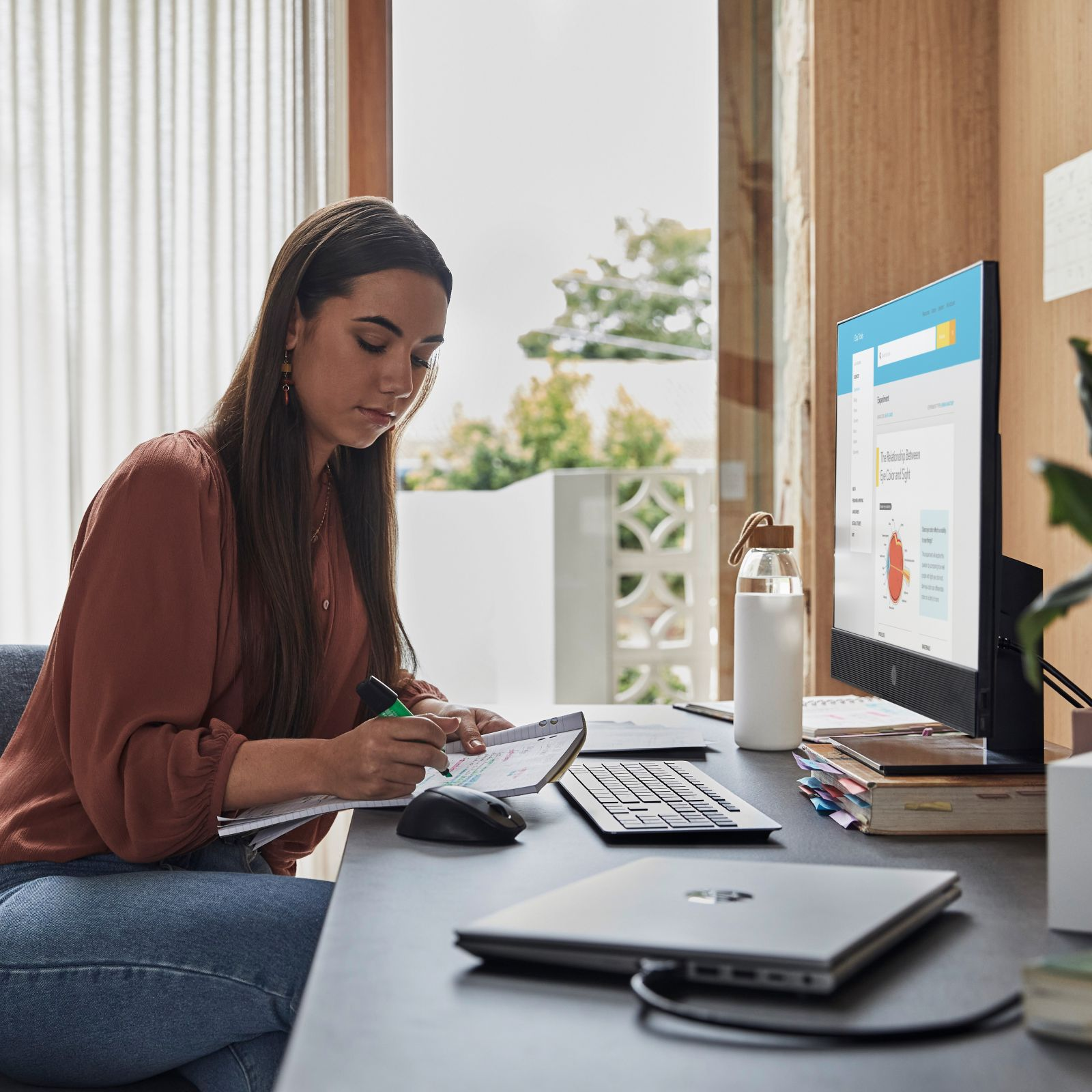 Kobieta pisząca na notatniku przy biurku z komputerem, butelką wody i klawiaturą w domowym biurze.