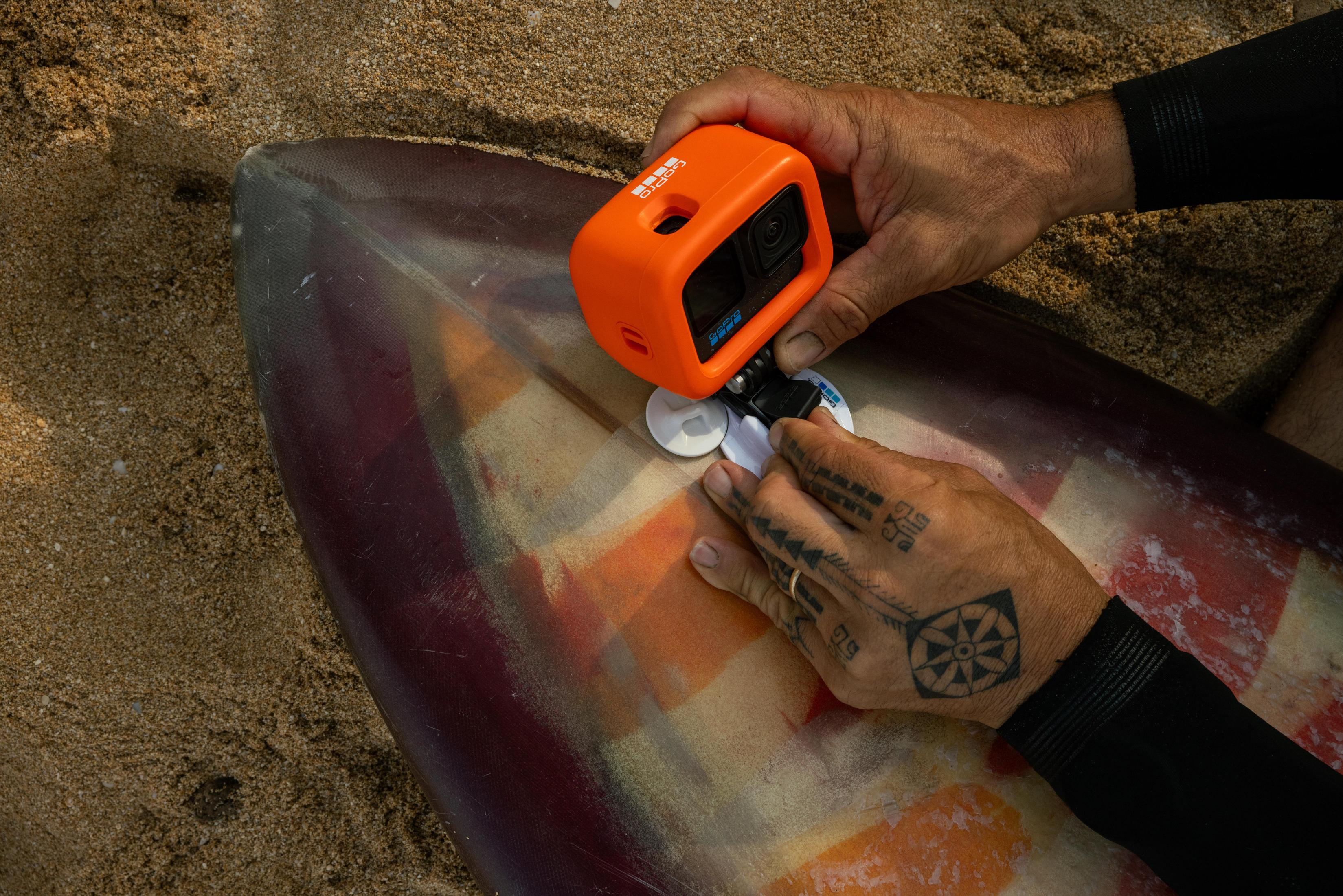 Person montiert eine orange GoPro-Kamera auf einem Surfbrett. Tätowierte Hände.