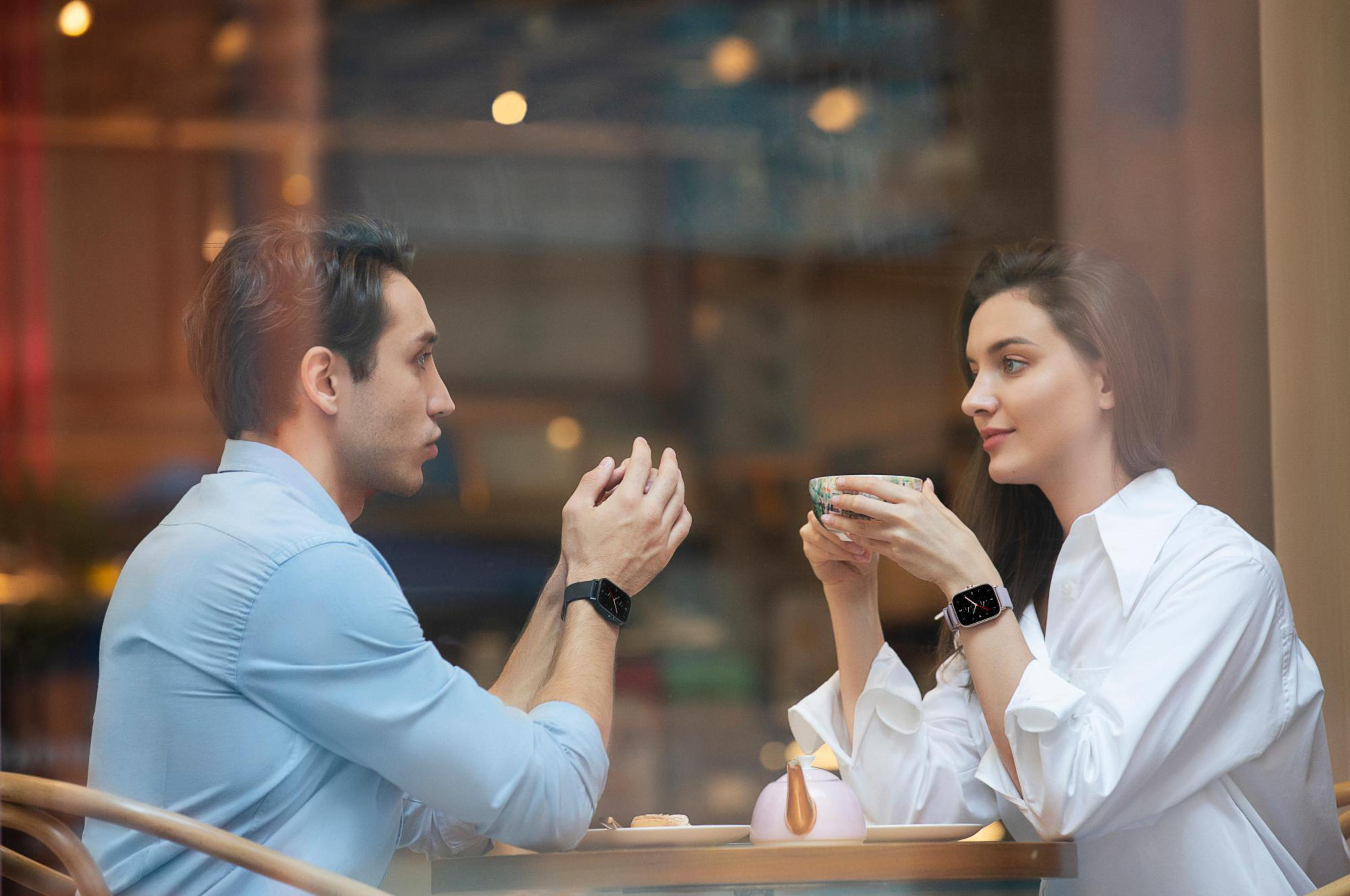 Coppia in un caffè, con smartwatch. Si guardano, parlano. Teiera sul tavolo.