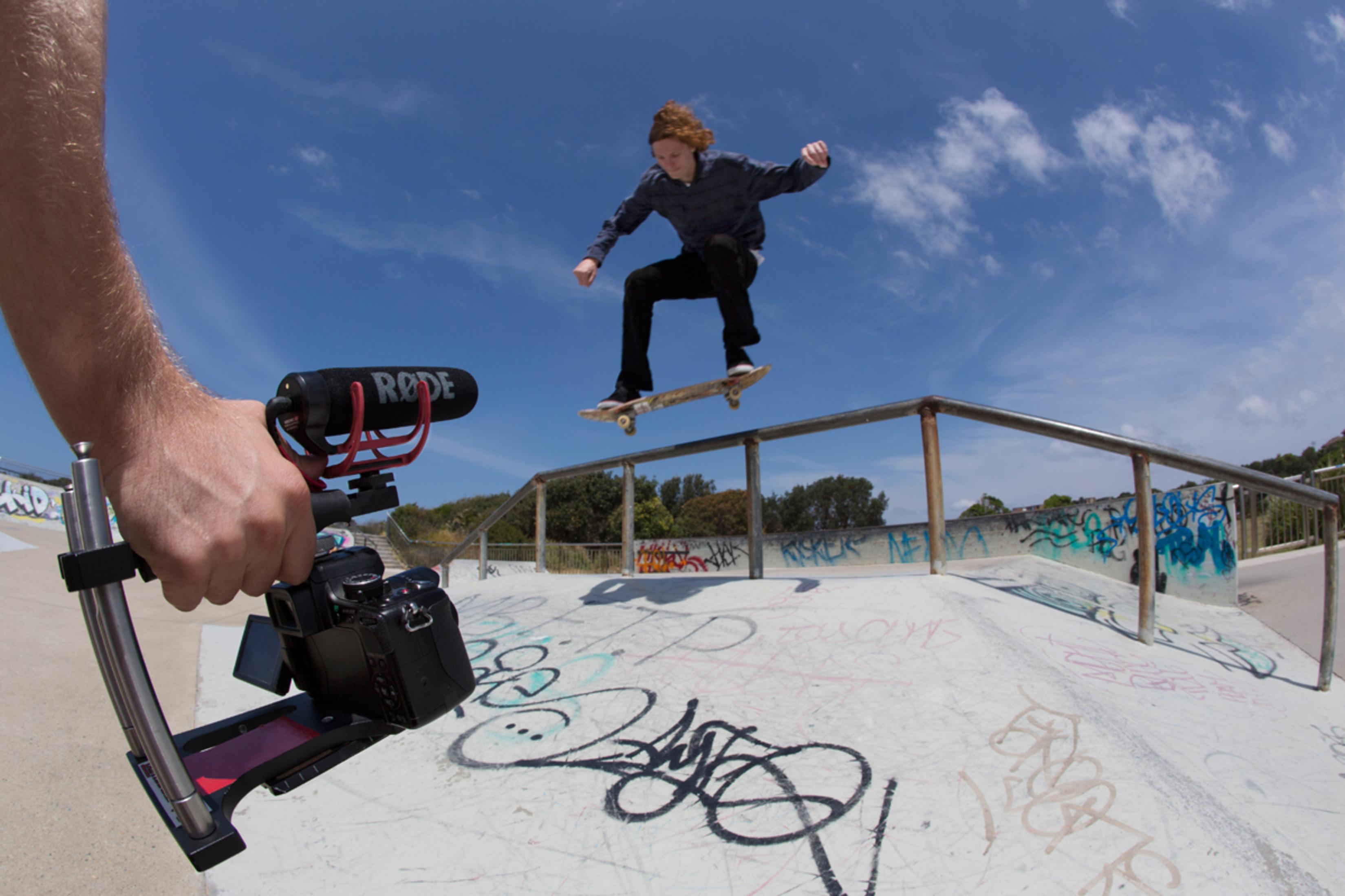 Osoba filmująca deskorolkarza wykonującego trik w skateparku za pomocą kamery i mikrofonu.