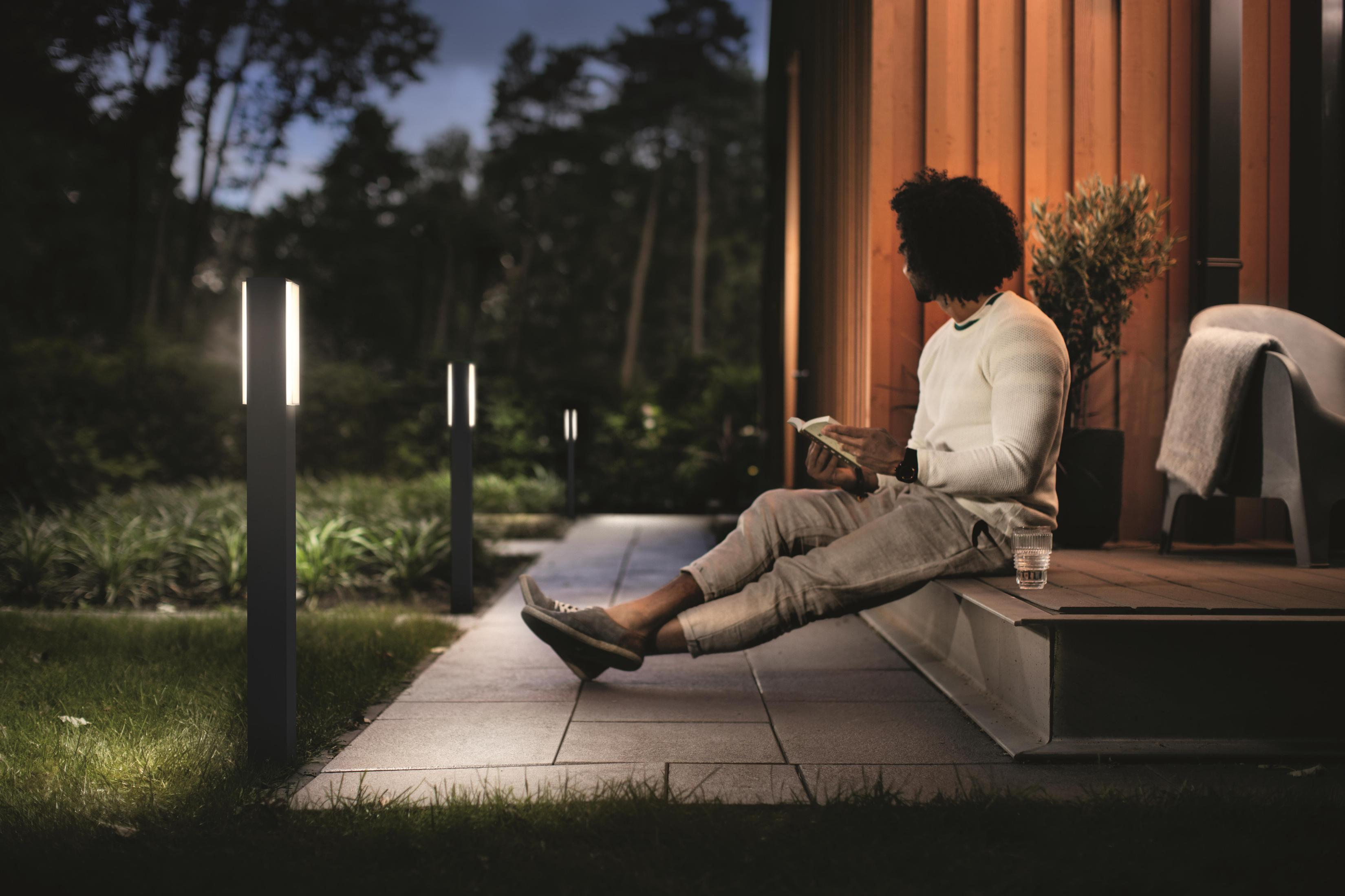 Ein Mann liest abends auf einer Veranda ein Buch, mit Lichtern, einem Stuhl und einem Wasserglas.