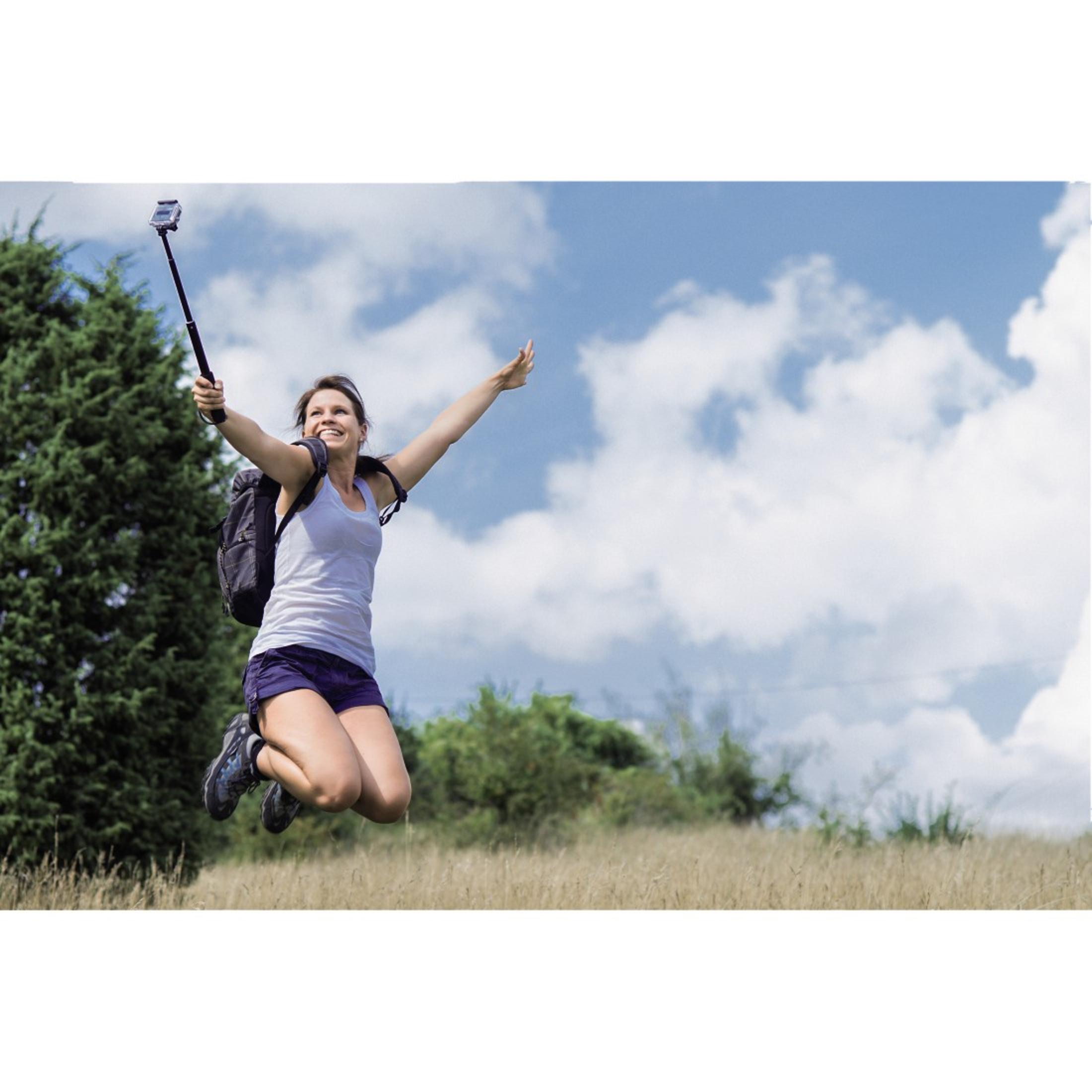Frau springt mit Selfie-Stick. Bäume und Himmel im Hintergrund. Glückliche Frau, Arme hoch.