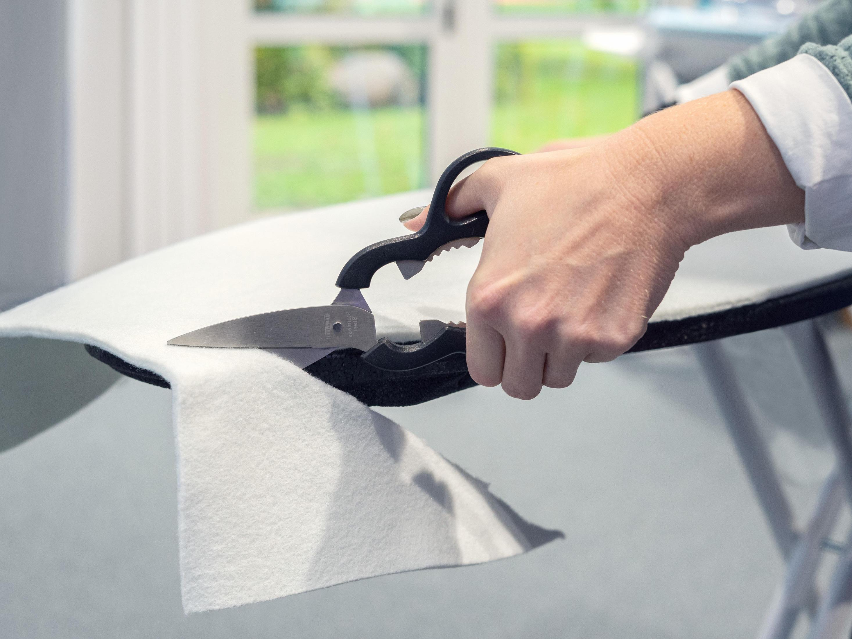 Dłoń używająca nożyczek do cięcia białej tkaniny na desce do prasowania.