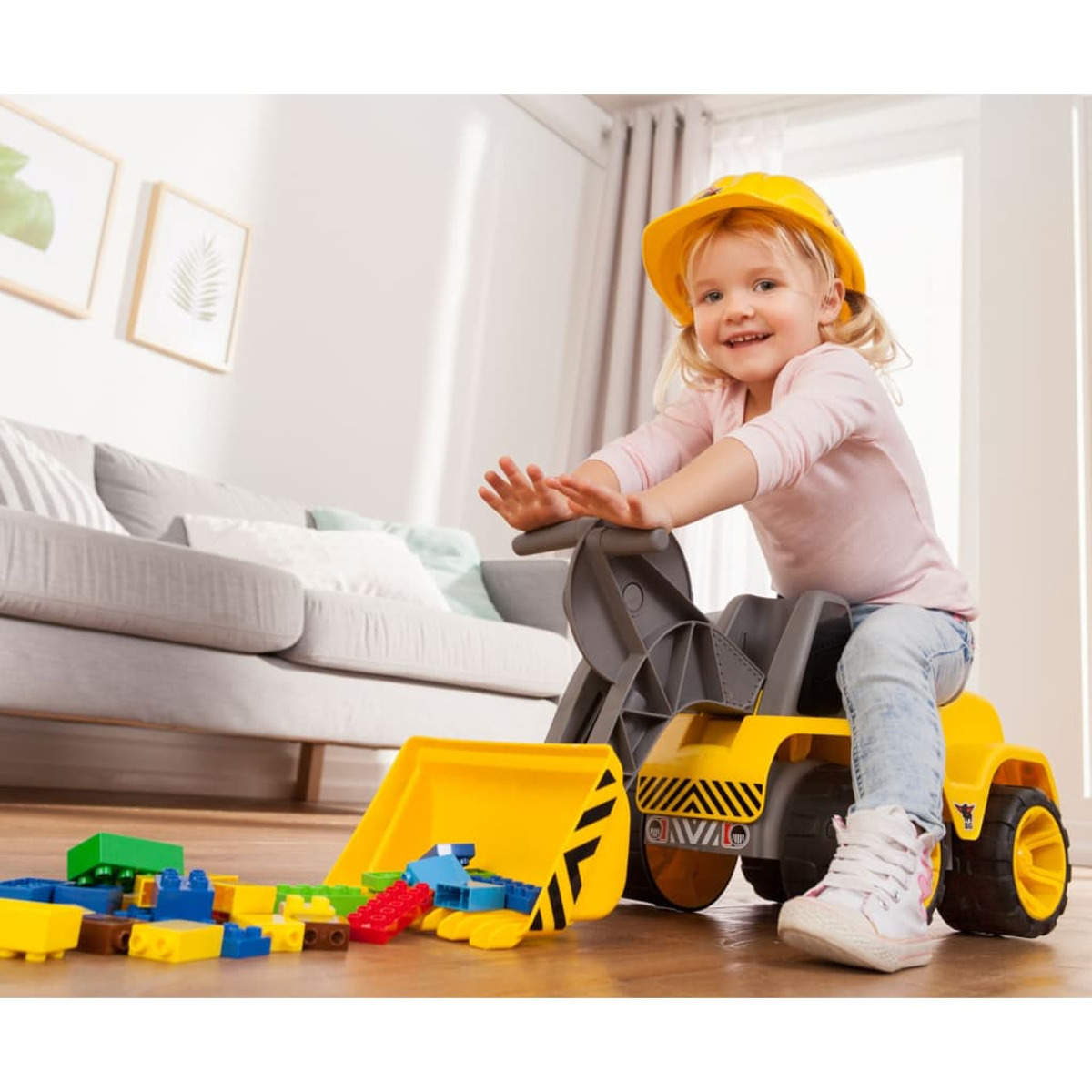 Dziecko w żółtym kasku siedzi na żółtej zabawce-koparce, otoczone klockami.