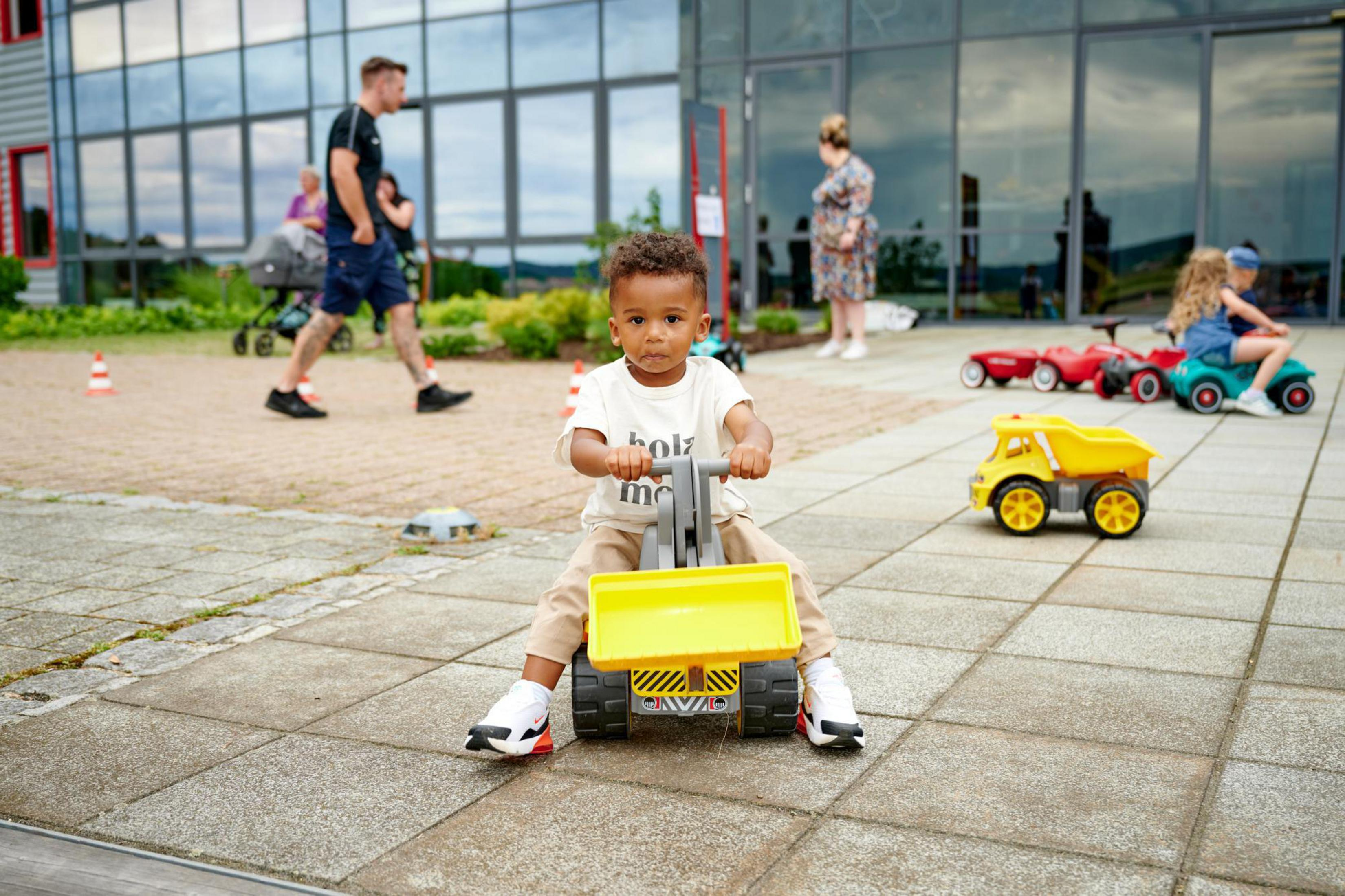 Małe dziecko na żółtej zabawce koparce, z innymi dziećmi, dorosłymi i budynkami.