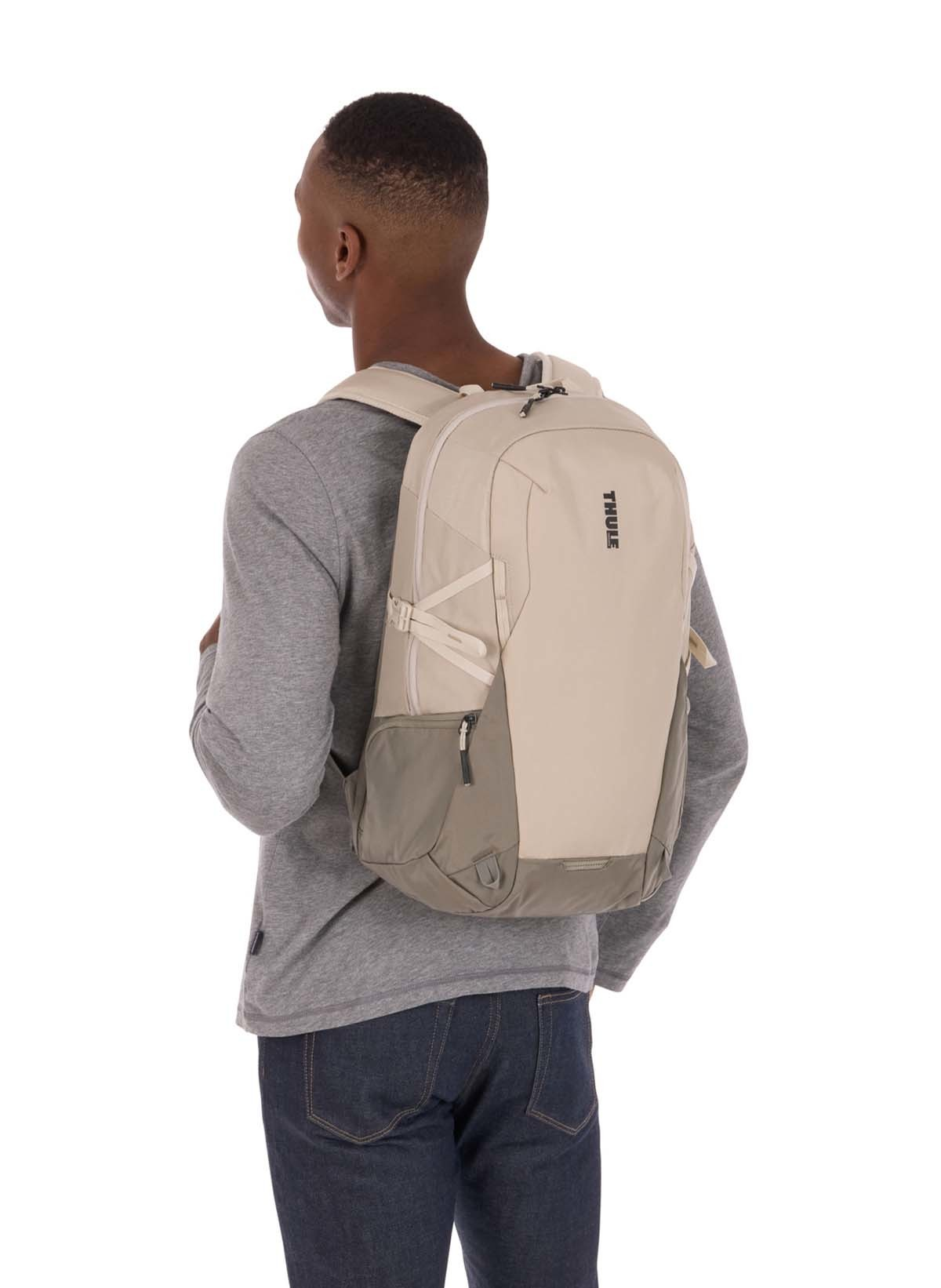 Ein Mann mit grauem Hemd und beigem Rucksack steht vor weißem Hintergrund, von hinten gesehen.