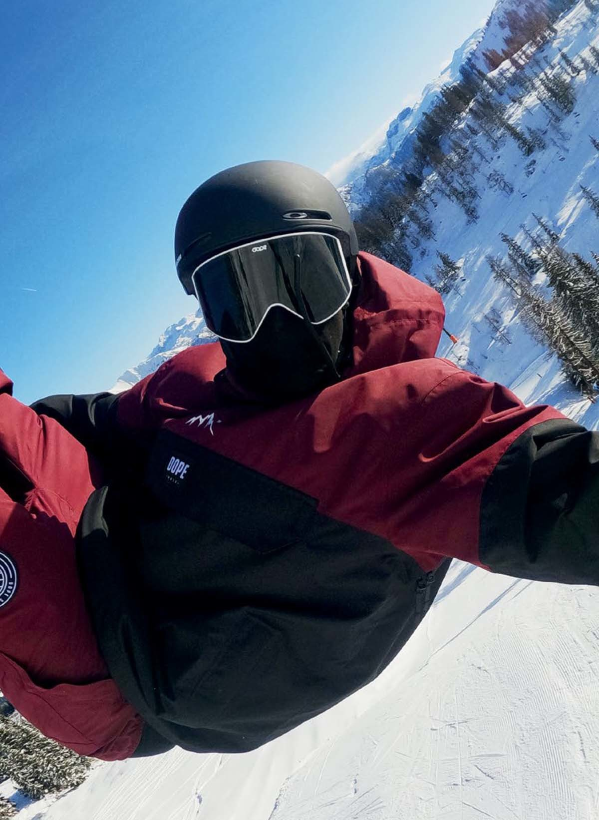 Un snowboarder en tenue noire et bordeaux est en l'air, devant un paysage de montagne enneigé.