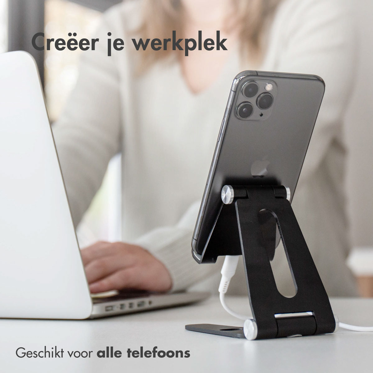Een persoon werkt op een laptop, met een telefoon op een zwarte standaard ernaast. Witte kabel aangesloten.