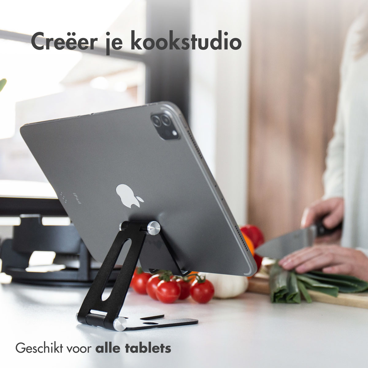 Tablet op een standaard, met keukenomgeving, tomaten en een persoon die snijdt.