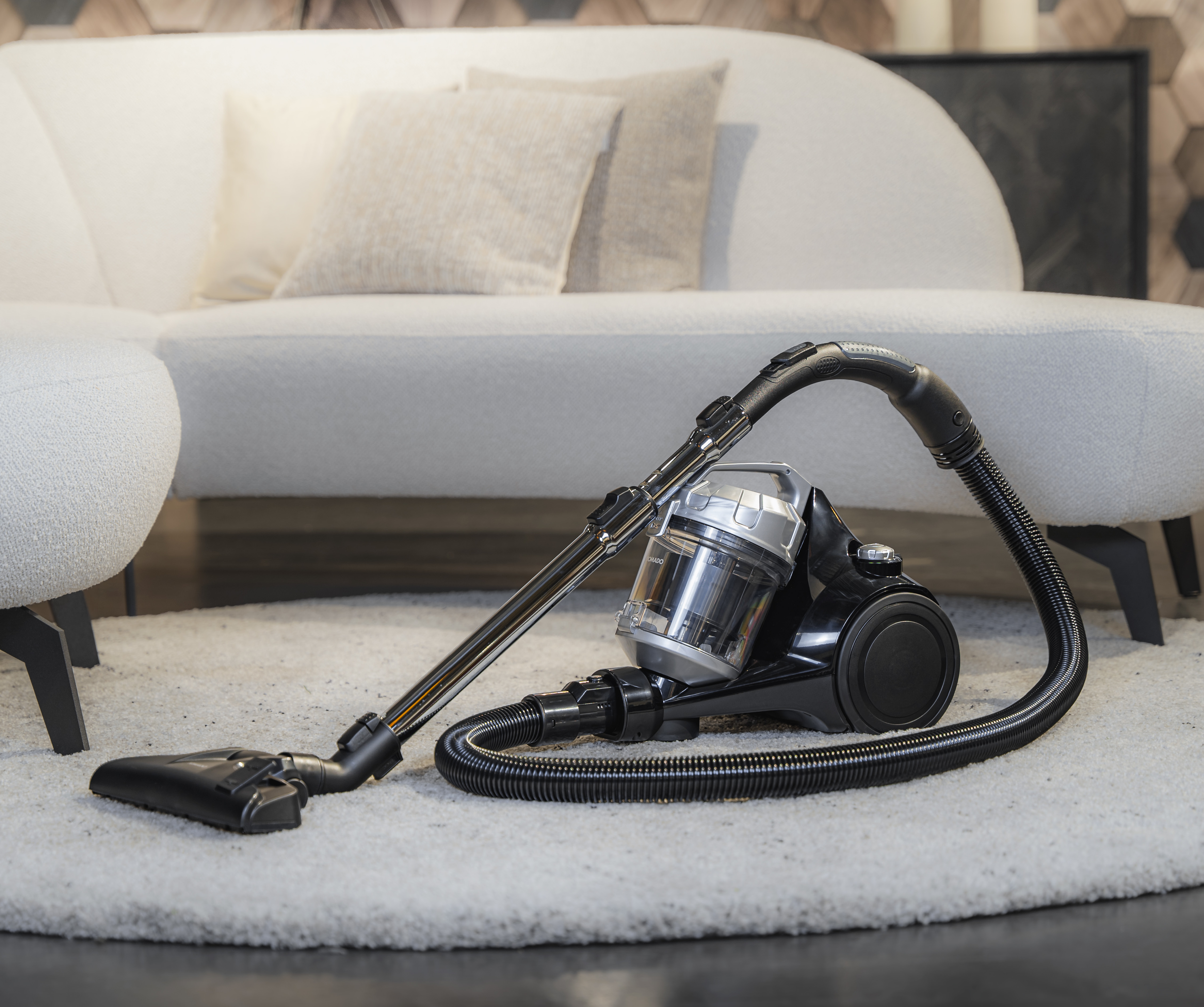 Black vacuum cleaner on rug, hose and attachments, white couch, light background.