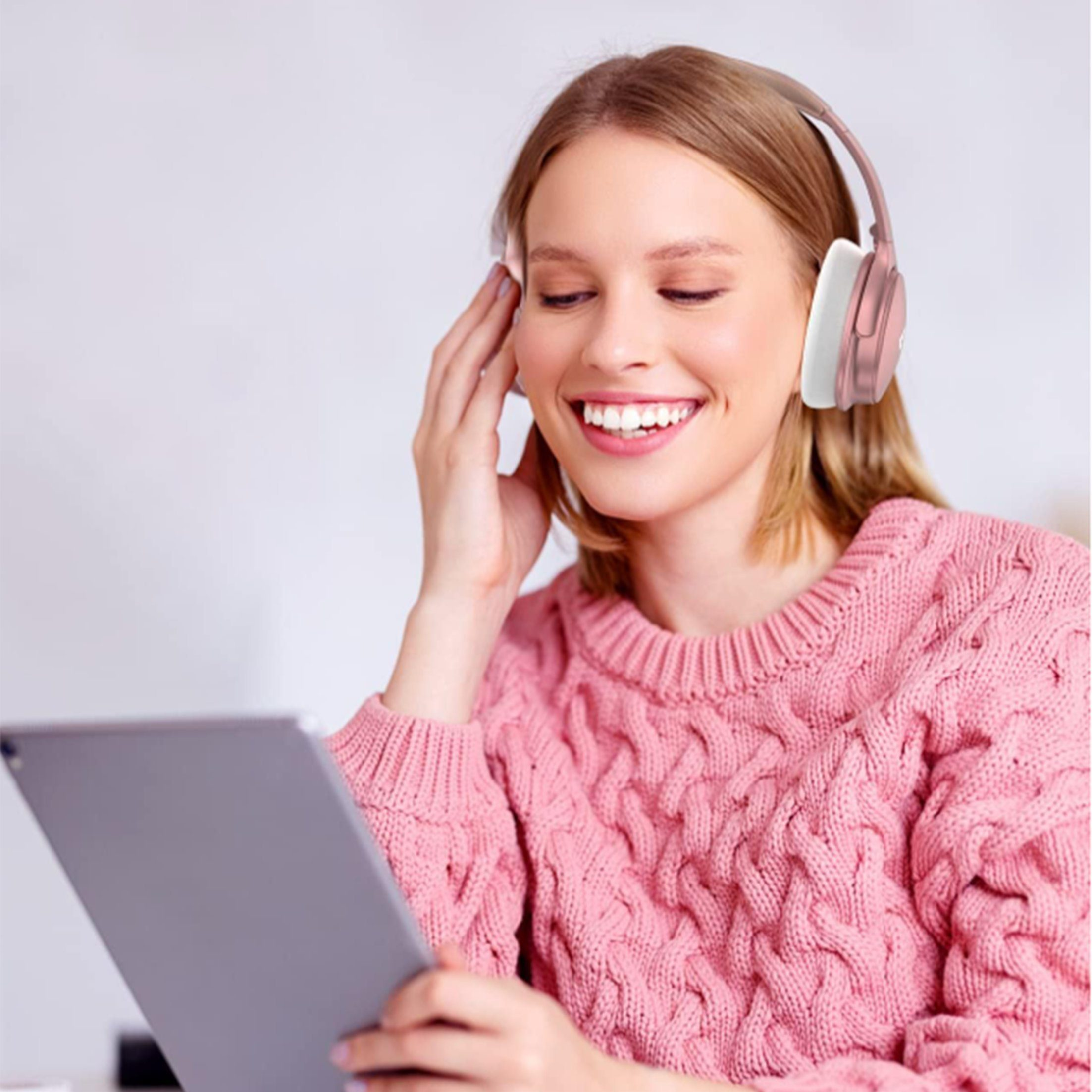 Eine Frau mit Kopfhörern und einem rosa Pullover lächelt, während sie ein Tablet hält. Der Hintergrund ist weiß.
