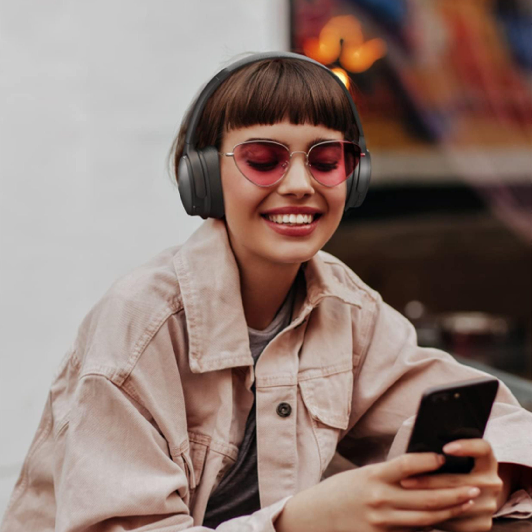 Frau mit Kopfhörern und rosa Sonnenbrille, hält ein Telefon. Lächelt.