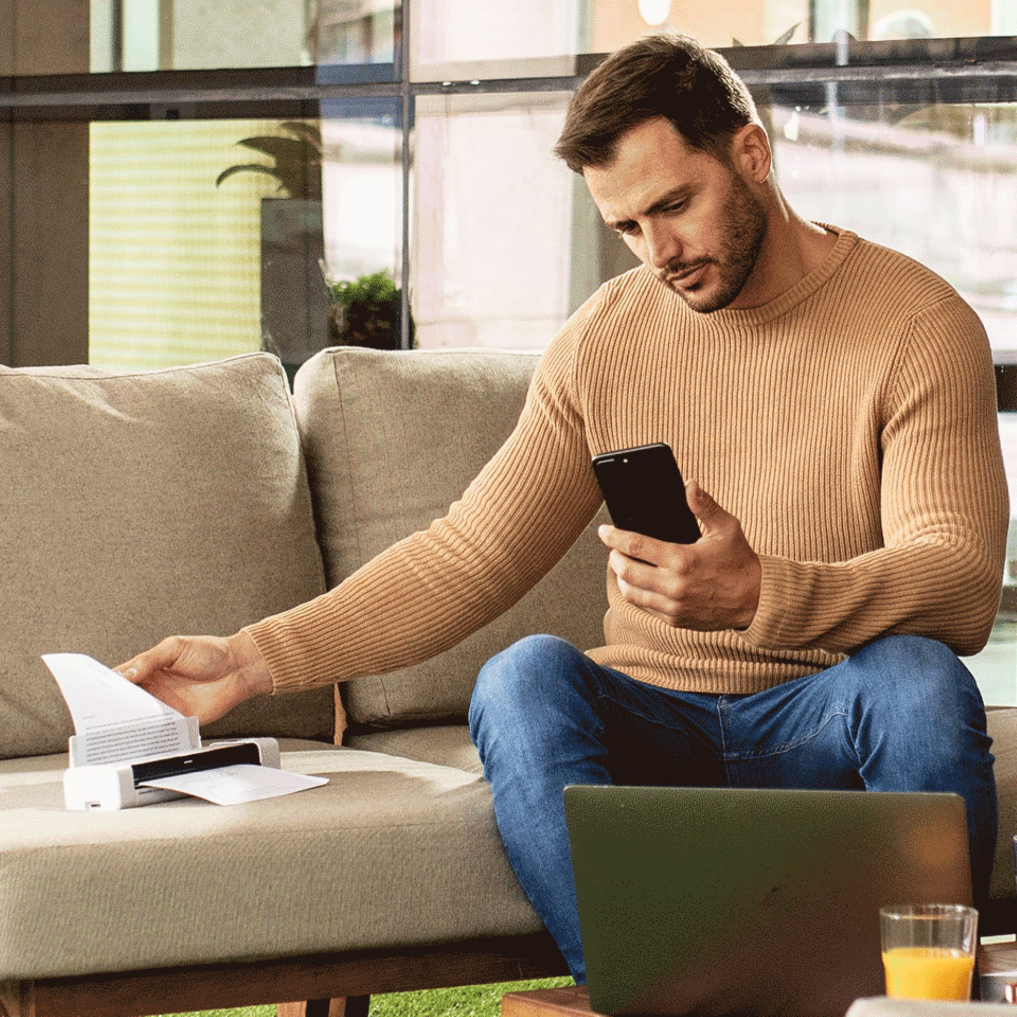Mann sitzt, schaut auf Telefon, hält Papier, in der Nähe eines Druckers und Laptops. Sofa Hintergrund.
