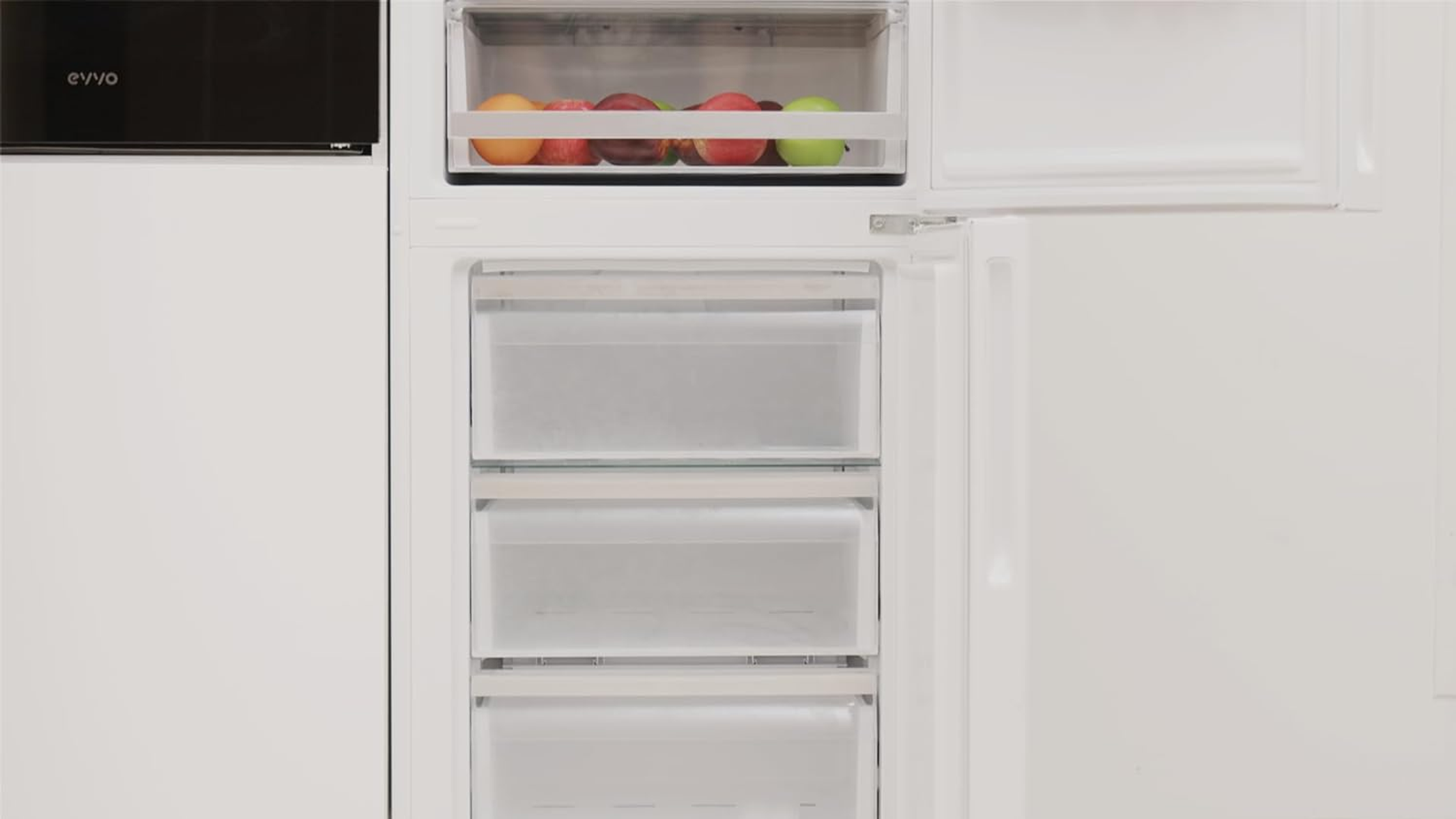 Refrigerador blanco con cajones de congelador abiertos, con frutas. Interior blanco con panel negro.